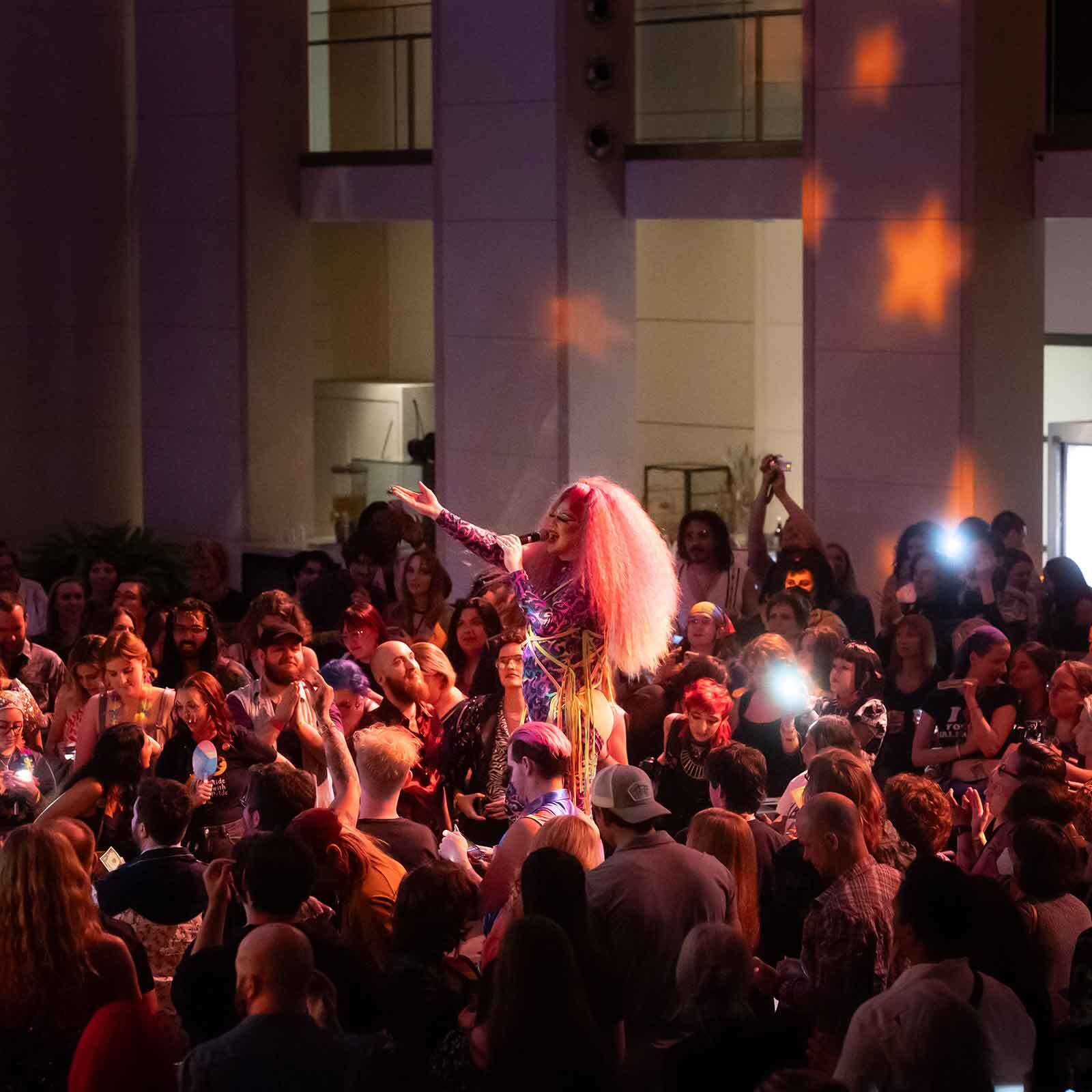Performer with pink hair sings into a microphone in a crowded indoor audience under warm lighting.