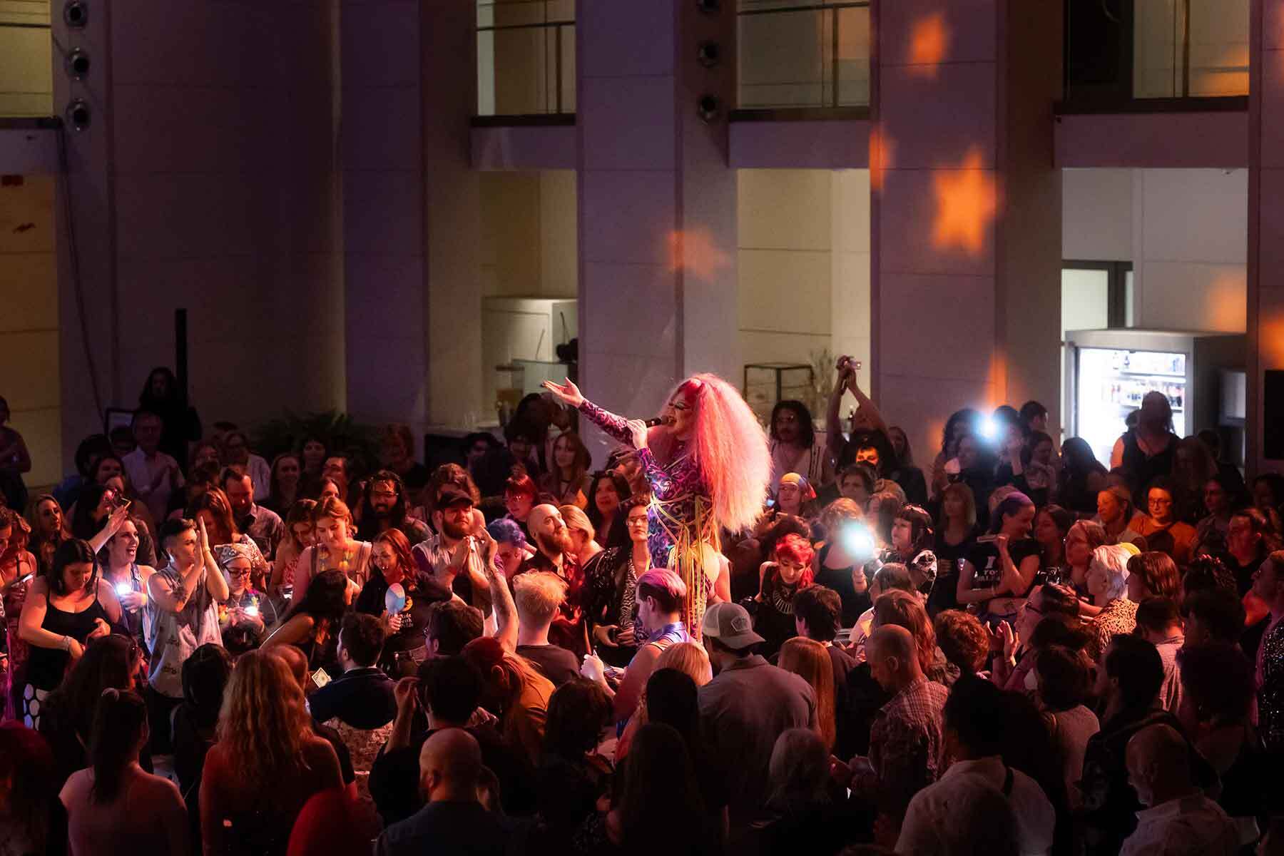 Performer with pink hair sings into a microphone in a crowded indoor audience under warm lighting.