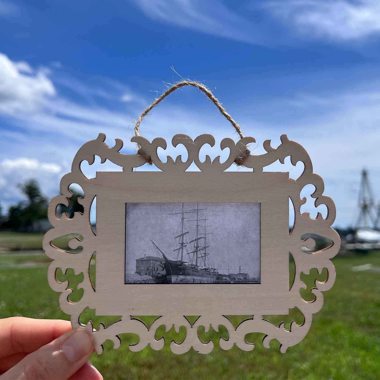 A hand holds an ornate wooden frame containing a historic black-and-white photograph of a tall-masted ship, with a grassy waterfront and blue sky in the background.