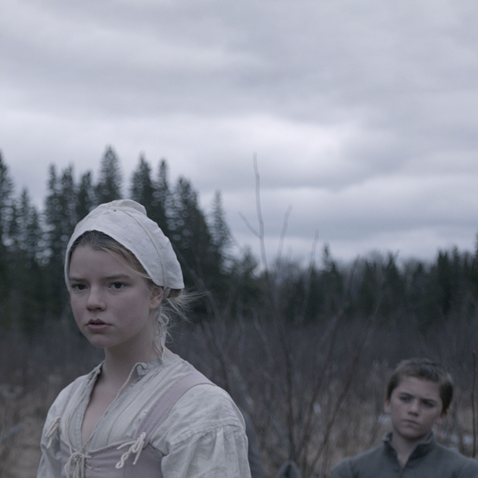 Scene from the Witch, depicting actors Anya Taylor-Joy and Harvey Scrimshaw in period clothing, standing in a forest.