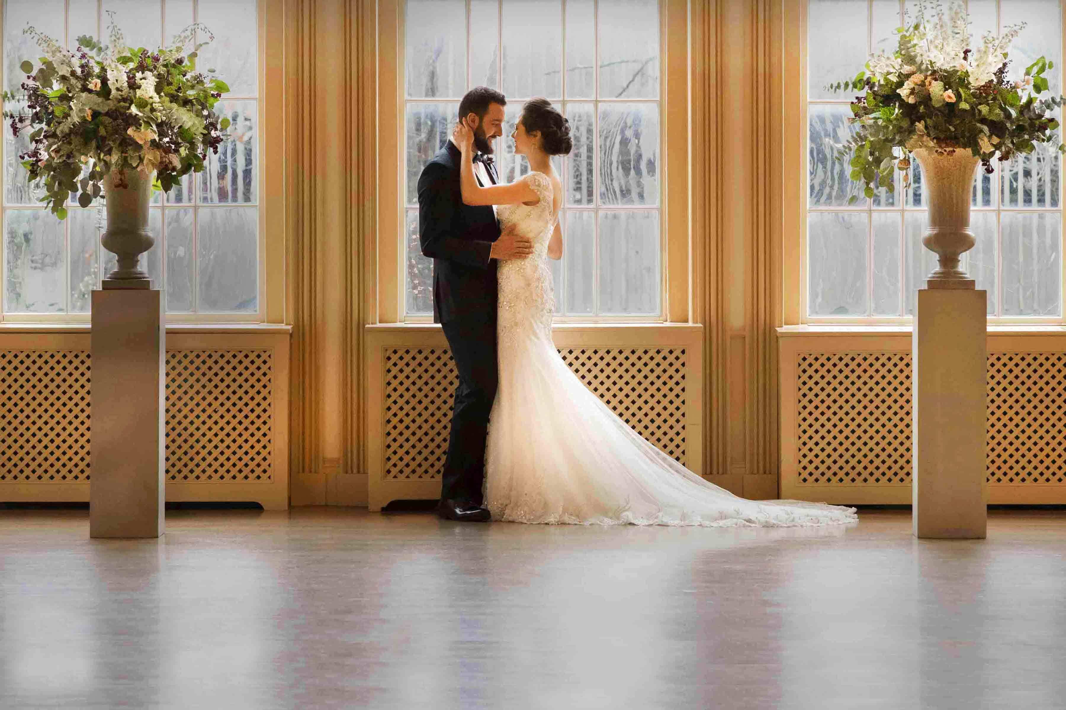 A bride and groom embrace in front of the windows in the East India Marine hall