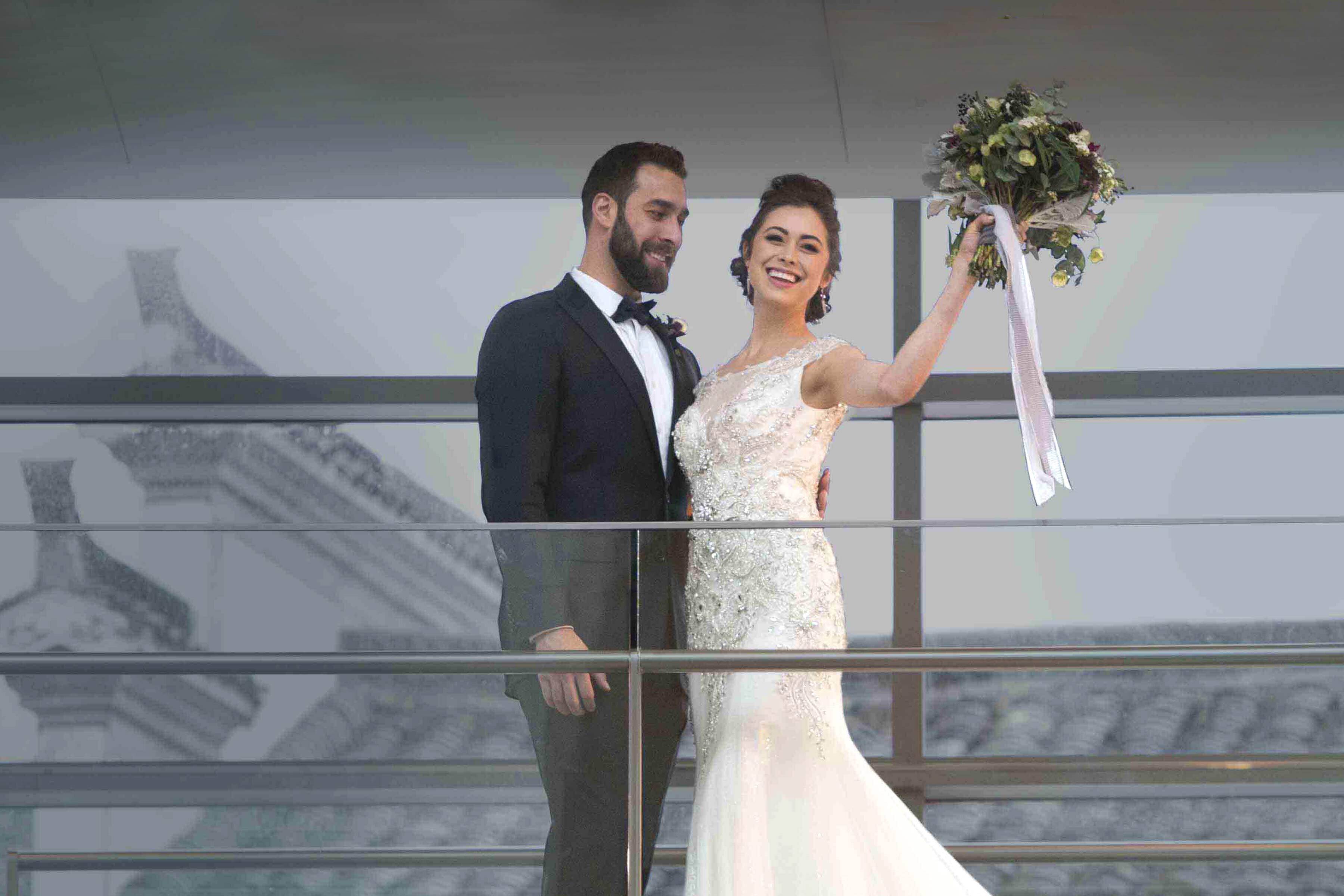 A bride holding up her bouquet stands with her groom on a balcony in PEM