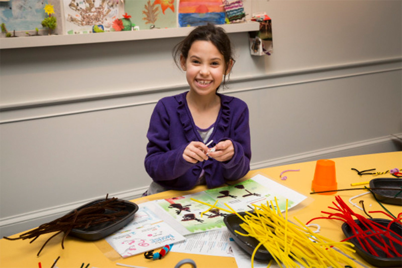 A smiling young girl with dark hair, wearing a purple sweater, sits at a yellow table filled with colorful craft supplies