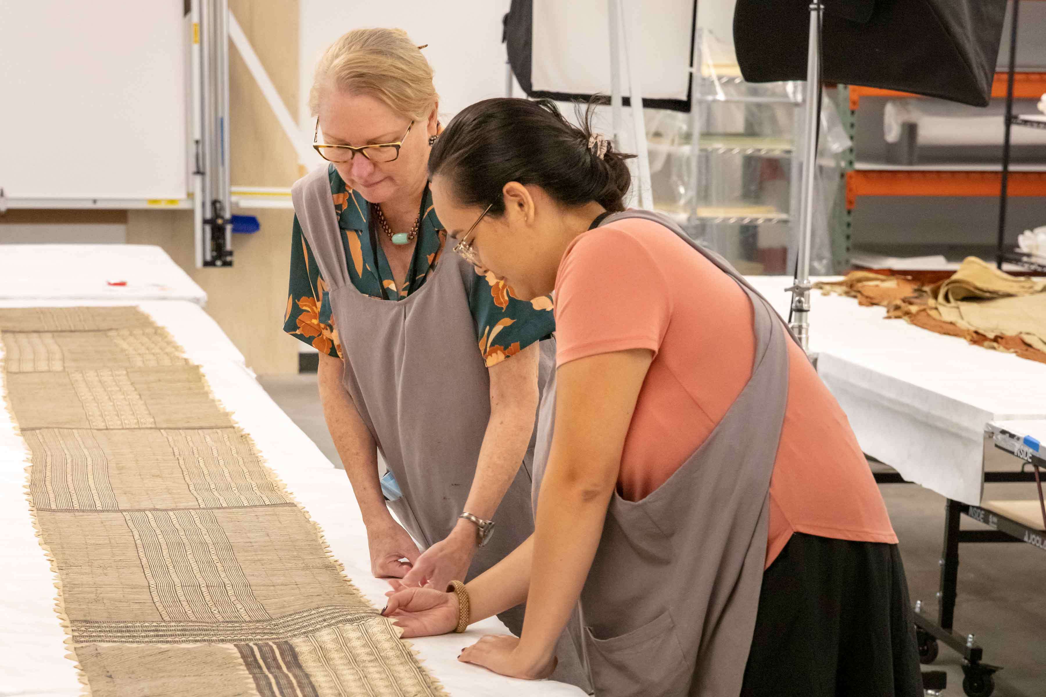 Two women working at a long white teble looking at a long woven textile