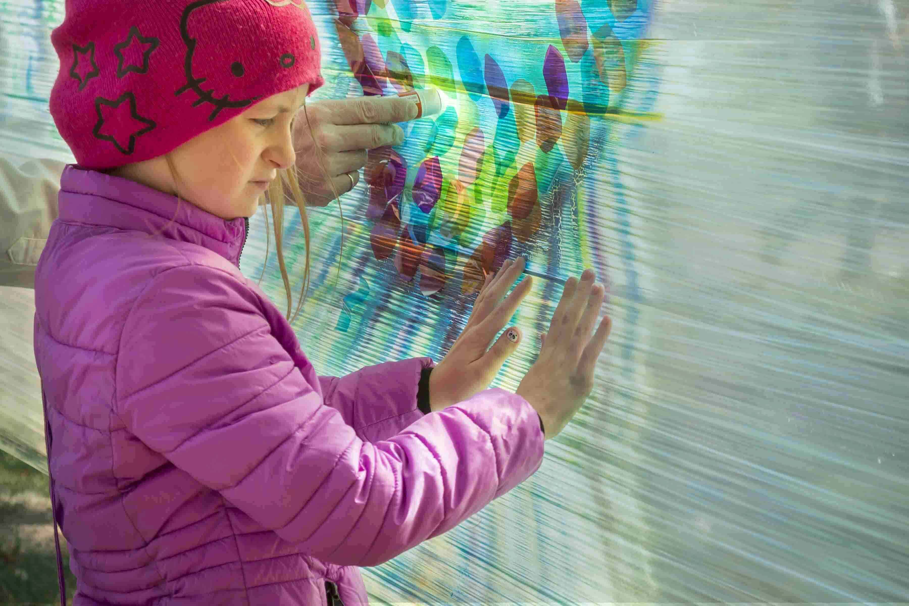 A young girl in a pink hat and purple jacket using her hands to paint onto a hanging fabric.