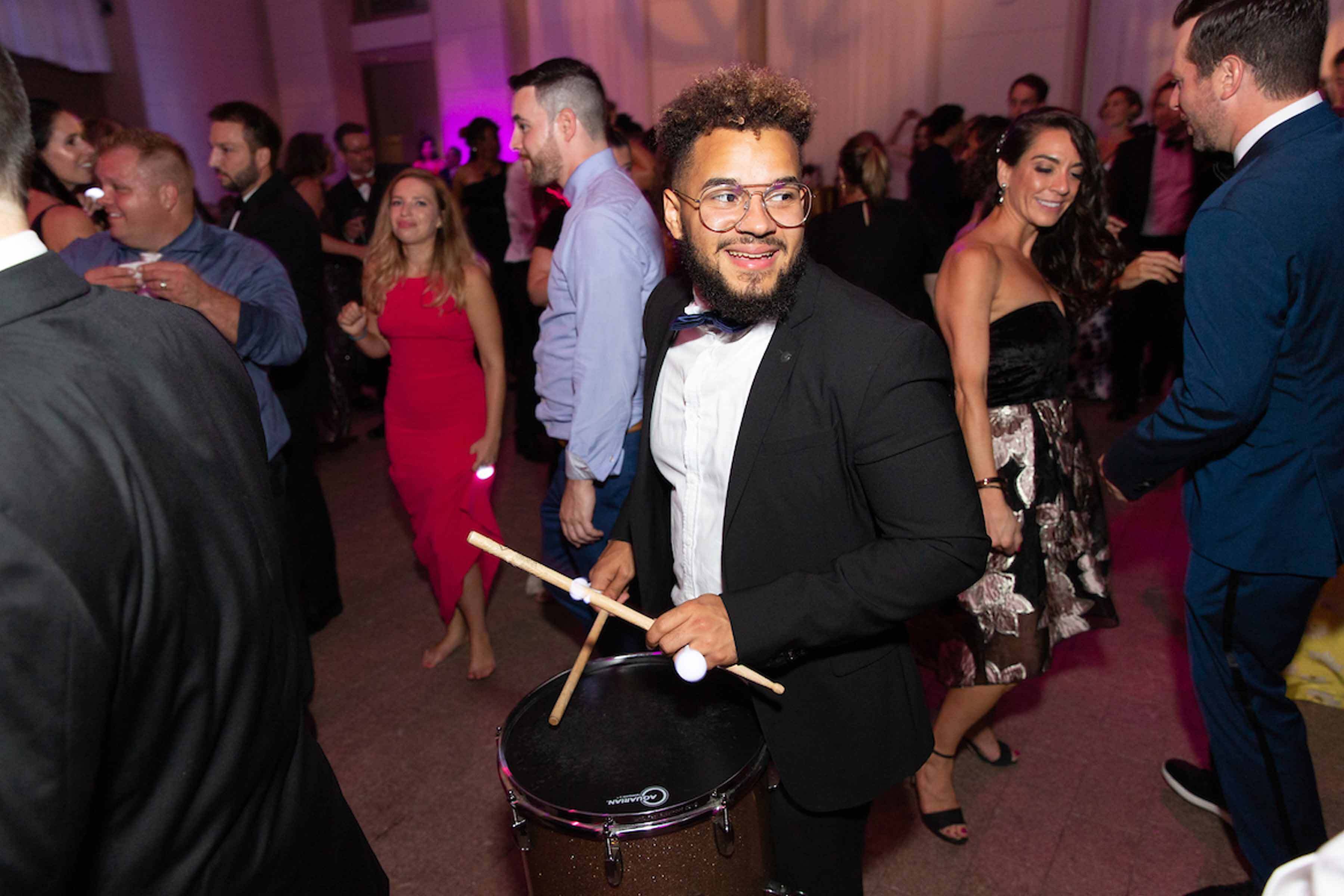 A man druming on a drum as he walks through a crowd of people during an event in a gallery