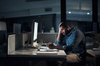 Someone working in the dark on a computer, looking at their phone seriously