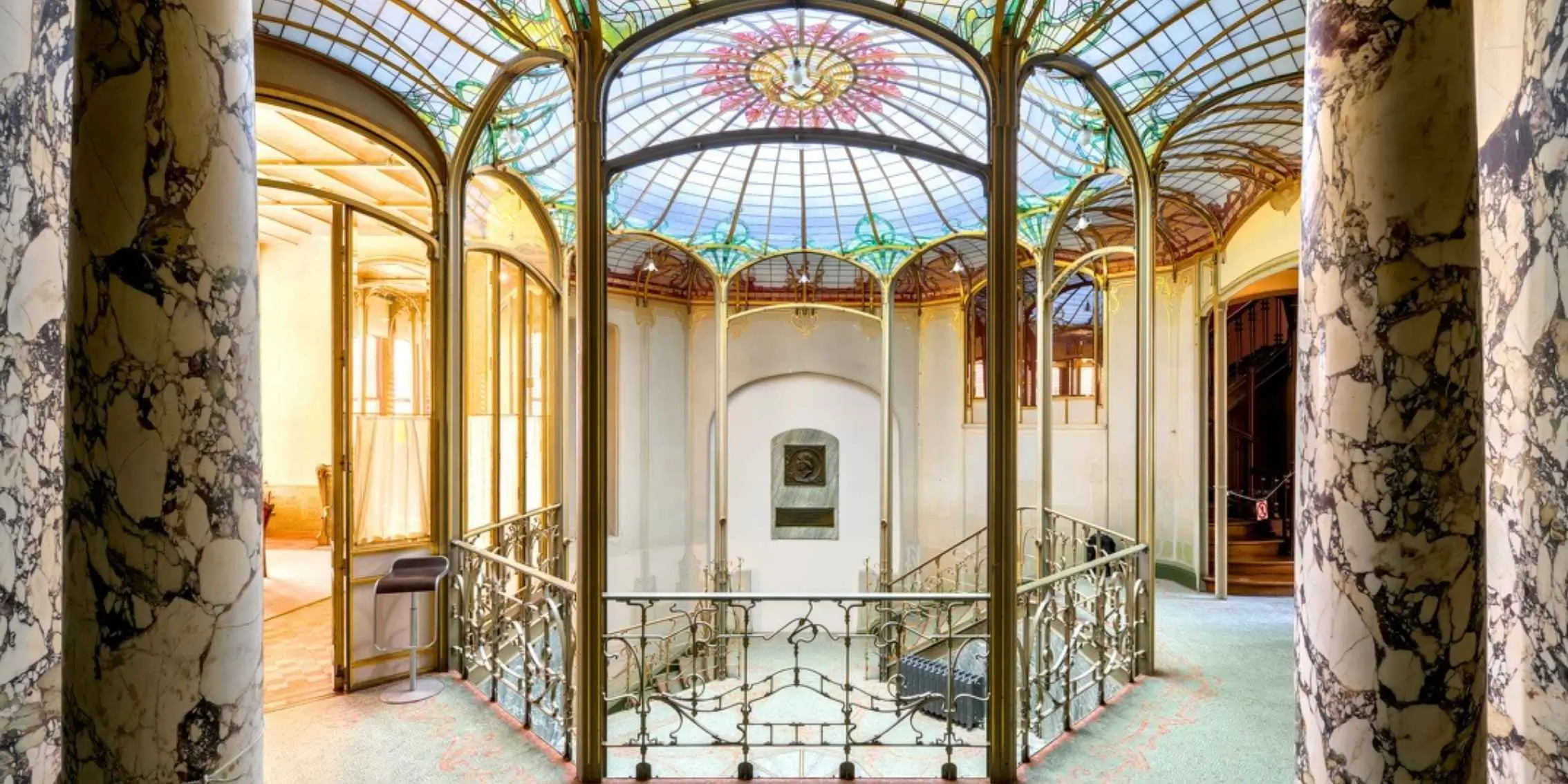 Interior of an Art Nouveau building with marble columns, decorative wrought
    iron railings, and a stained glass domed ceiling