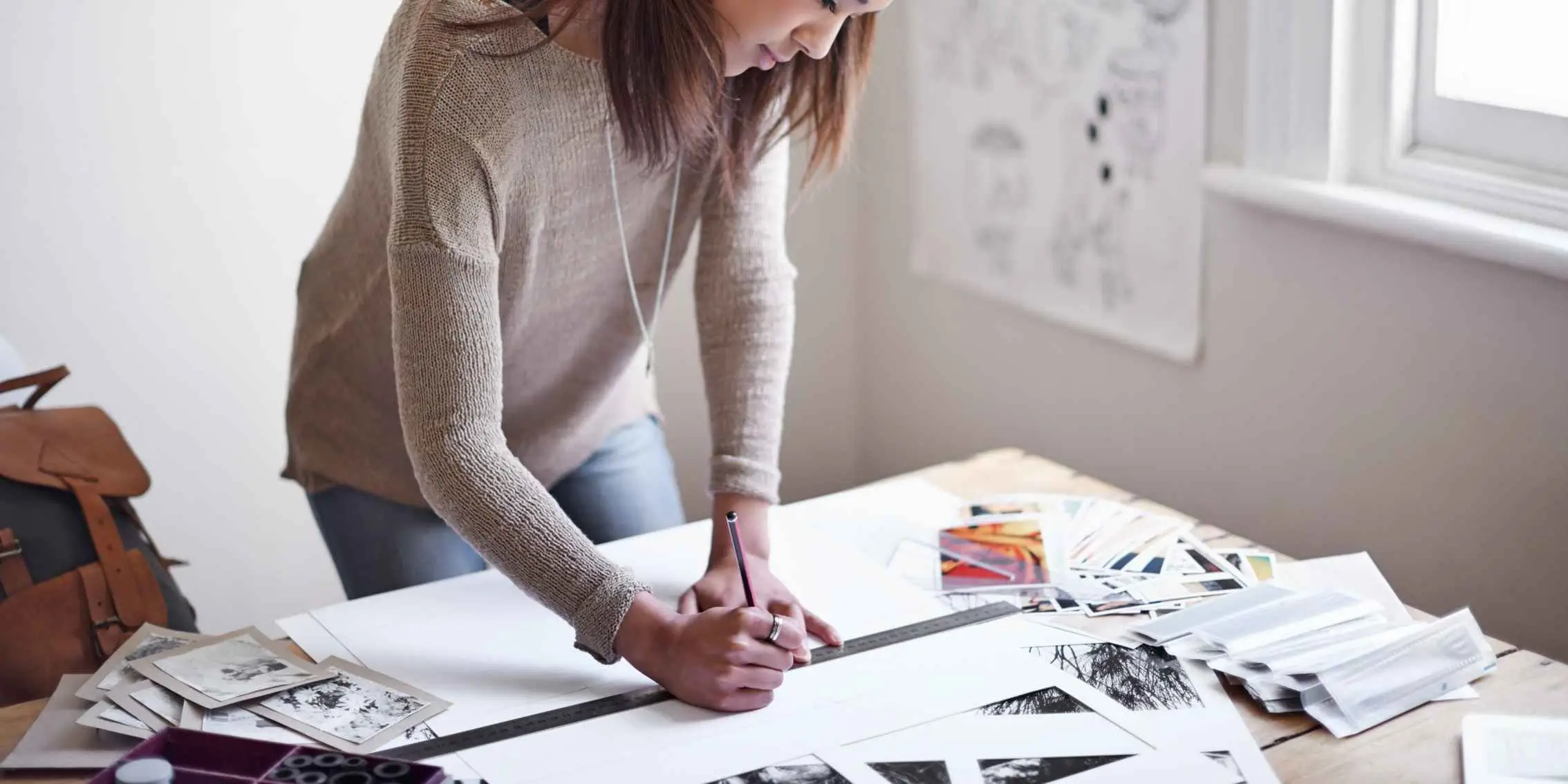 Person sketching on paper surrounded by black-and-white and color prints,
                working under natural light
