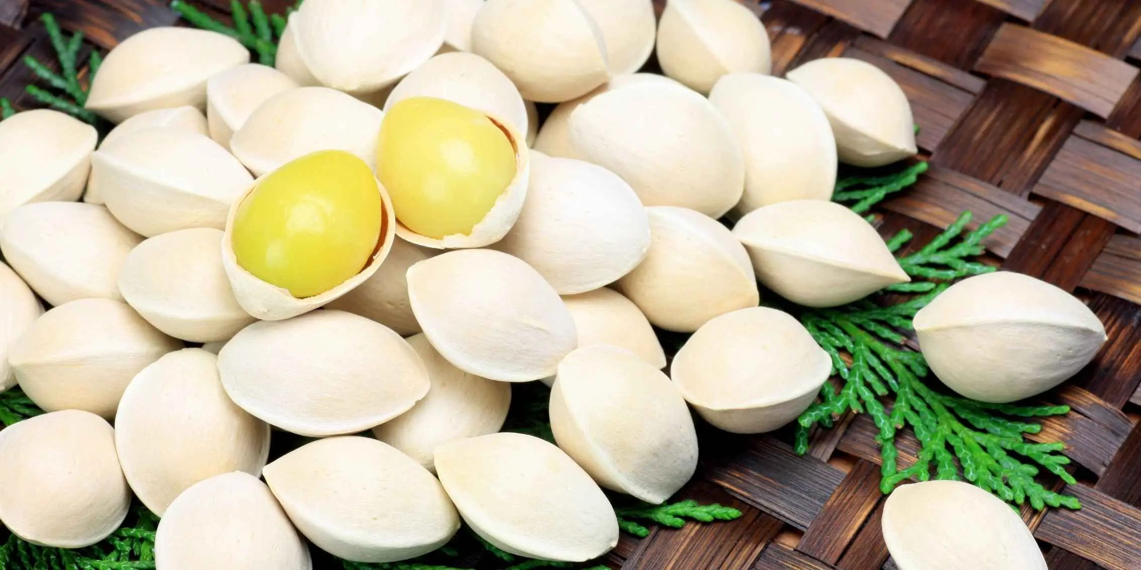 Whole ginkgo nuts with a few opened to reveal yellow seeds, displayed on a
    woven tray with green foliage