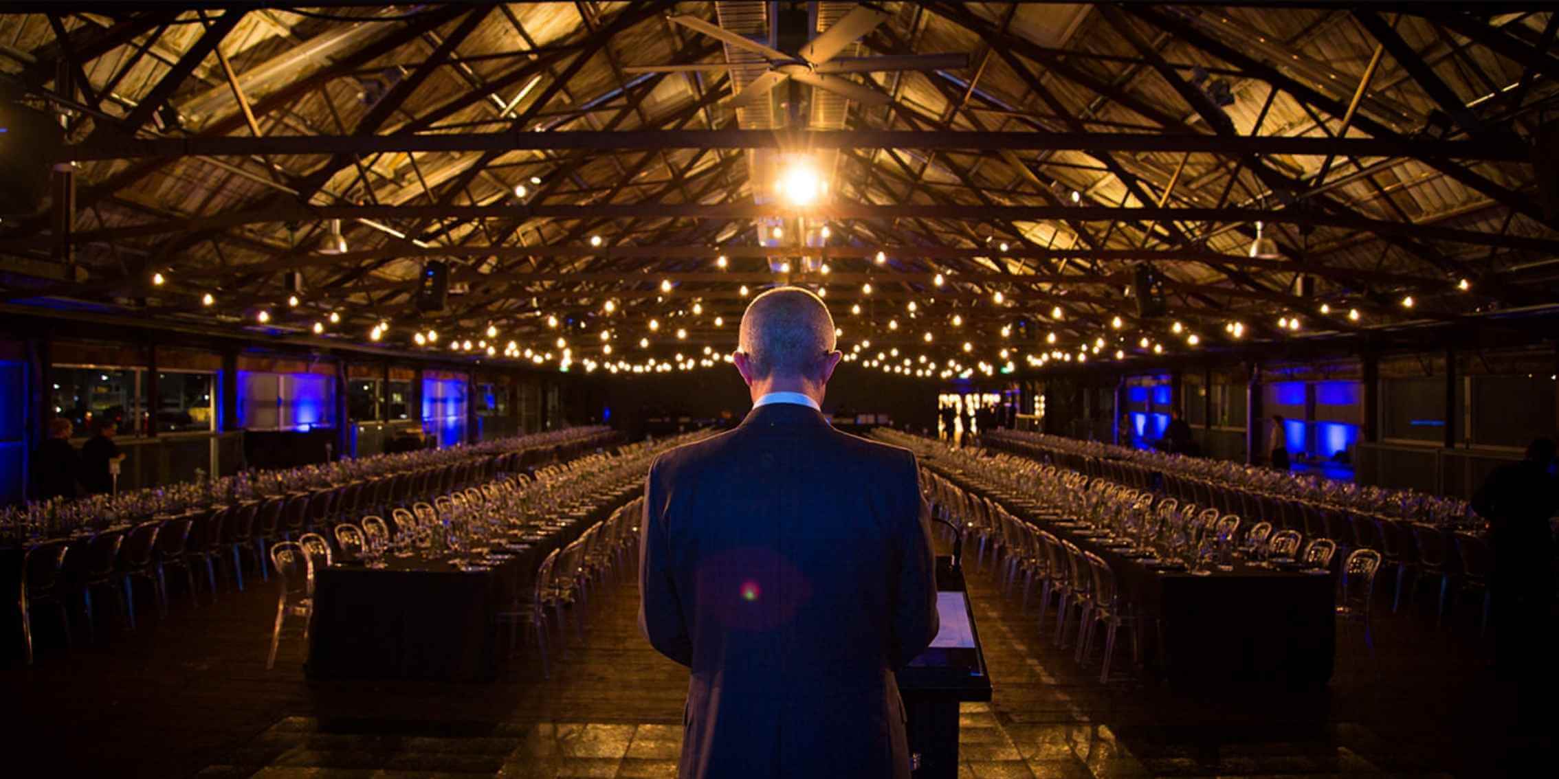 Why Photojournalism Matters Now More Than Ever – Capturing Moments of
Influence and Leadership A dramatic backlit shot of a speaker standing at a podium in a
dimly lit event hall, illuminated by string lights and ambient blue lighting.