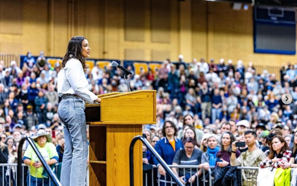Alexandria Ocasio-Cortez speaking at a wooden podium, wearing a long sleeve white shirt and blue jeans. She's angled to the right, looking out to a large crowd of people.