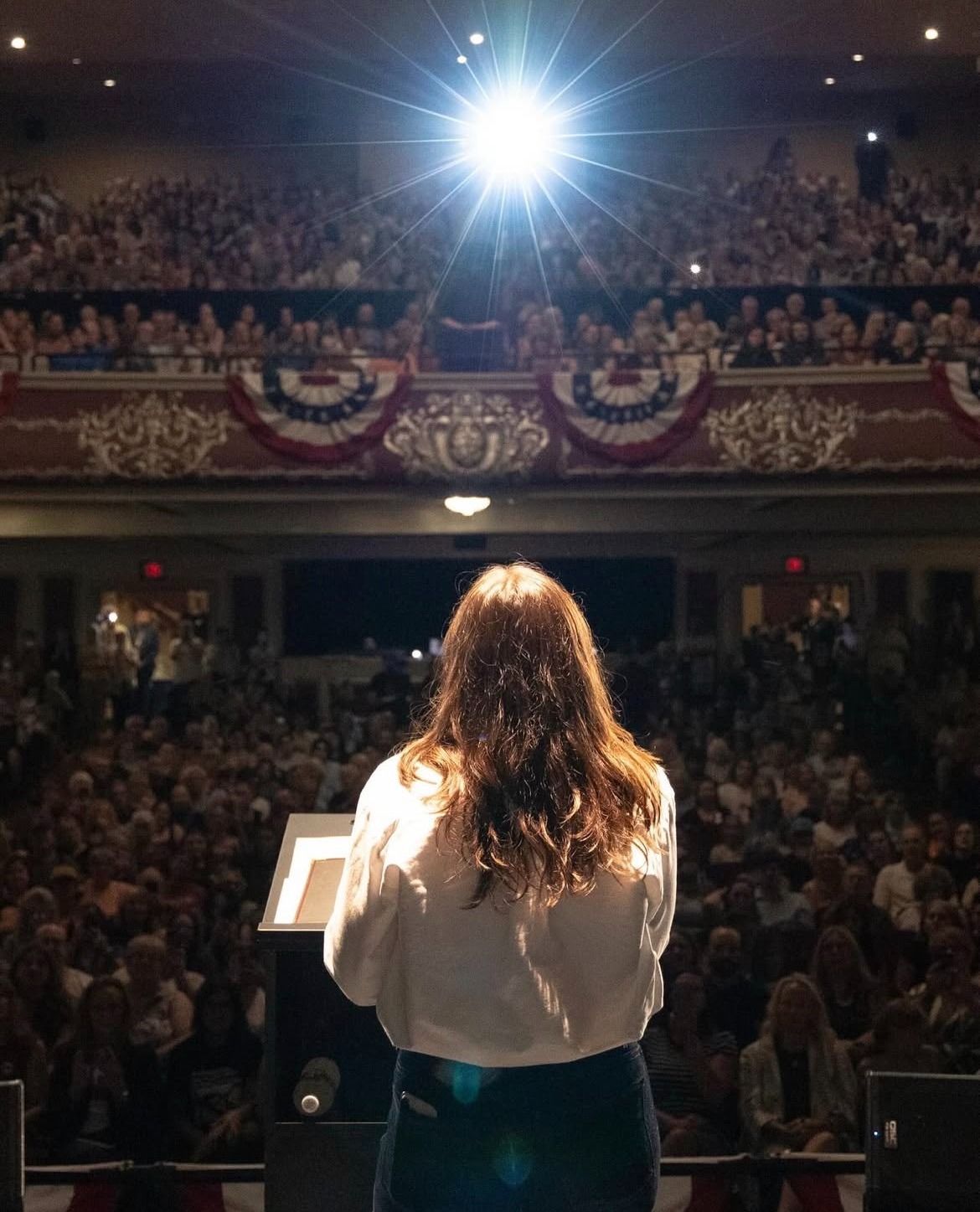 Photo taken from behind Alexandria Ocasio-Cortez as she stands at a podium addressing a full, two-tiered auditorium in Plattsburgh, New York.