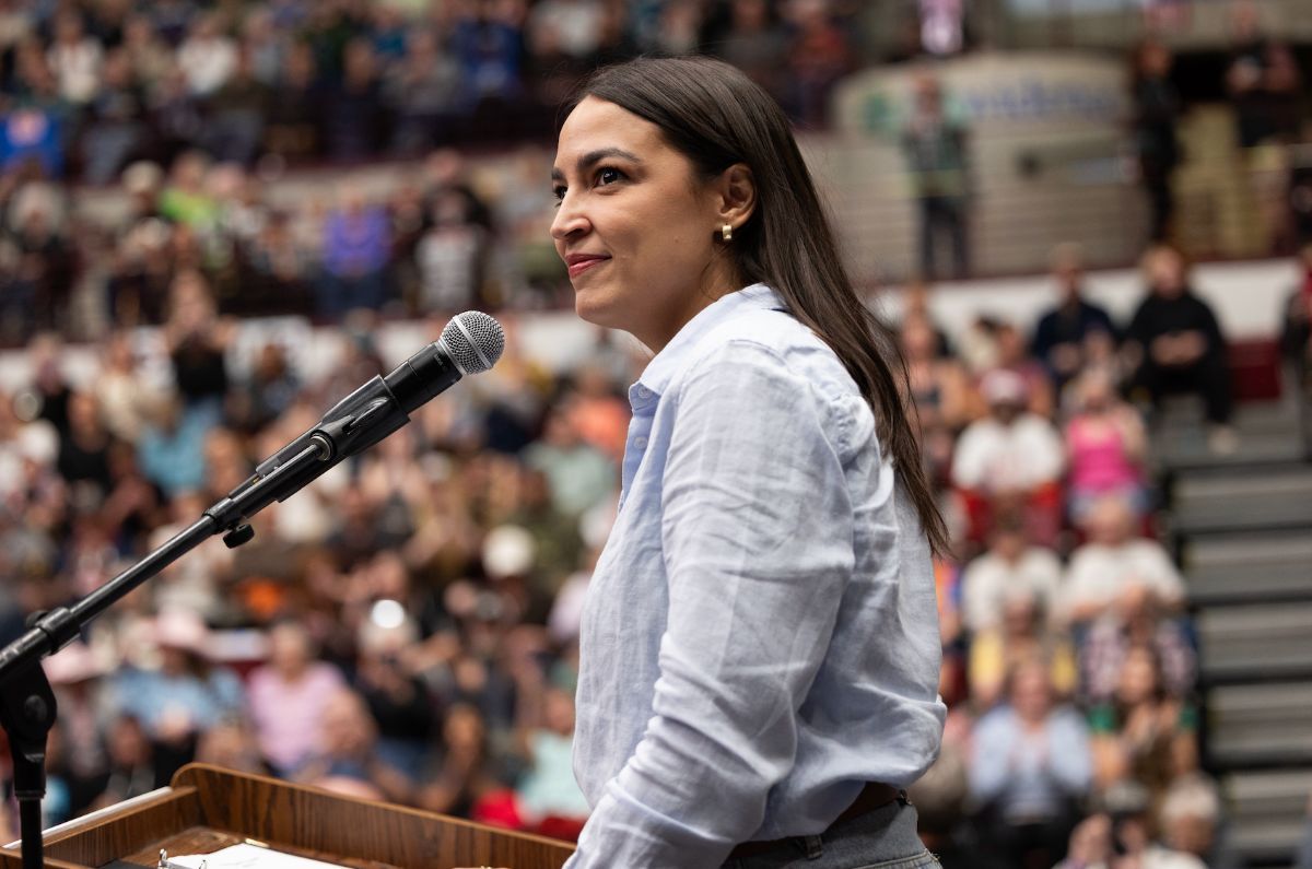 Alexandria standing at a podium with a microphone, her side facing the camera. She's looking off into the crowd slightly to her left, with a large crowd in the background