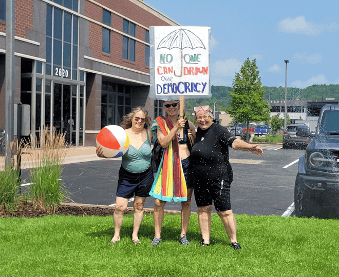 Photo of three Indivisibles in swimsuits, beach towels, and swim goggles outside a stuffy looking office park. One woman is holding a beach ball while the other holds a sign that says NO ONE CAN DROWN OUR DEMOCRACY