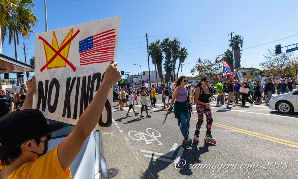 A protestor in California holds up a handmade NO KINGS sign while two women rollerblade past in the foreground and dozens of other No Kings protestors march in the background -- photo courtesy of zimmagery.com