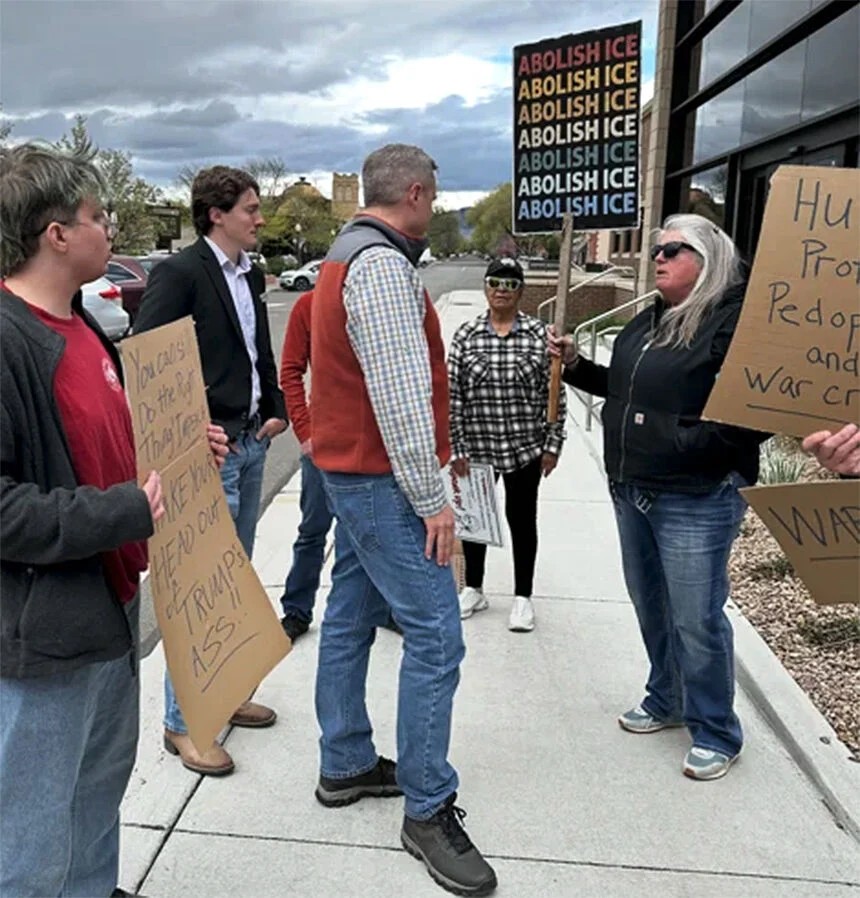 A small group of people standing on the sidewalk. Two of them are holding signs, one of which sayd ABOLISH ICE several times,
