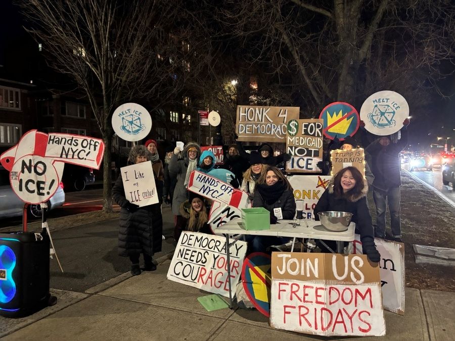 Several people sit at or stand behind a folding table set up on a street corner. The sun has already gone down and the people are wearing winter coats and hats, and holding or displaying many signs, including Melt ICE, $ for Medicaid not ICE, and a handpainted No Kings logo 