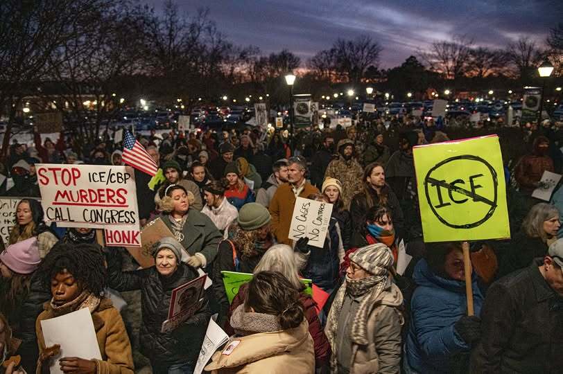 A large group of people, all dressed in winter coats, hats, scarves, etc, stand outside in the twilight. One is holding an American flag; many are holding signs. The signs we can read say STOP ICE/CBP MURDERS CALL CONGRESS HONK, NO CAGES NO CONTRACTS NO ICE, and ICE with a the sign for NO around it 