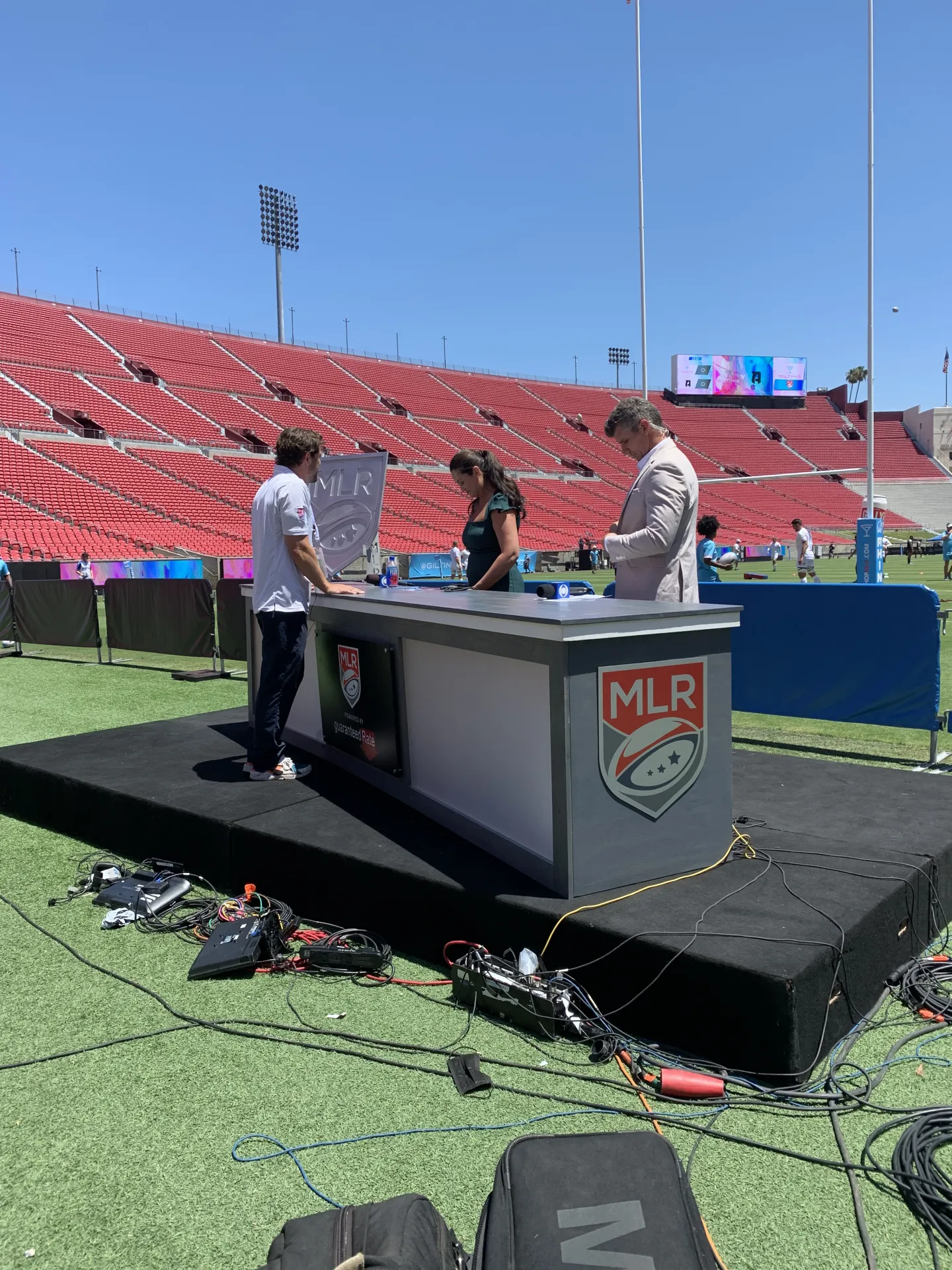 Outside on location setup of completed sportscaster desk with the crew setting up cables, lights, and surrounding signage.