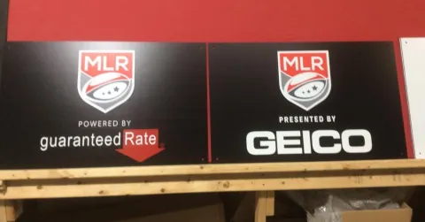 Completed black Major League Rugby sign with logo and Geico sponsor panel mounted across the front of a custom broadcast desk.