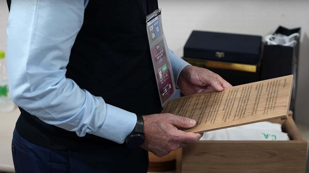 Ancelotti lendo carta de madeira Carlo Ancelotti de camisa azul e colete preto segura uma placa de madeira com texto gravado, lendo o presente no vestiário.