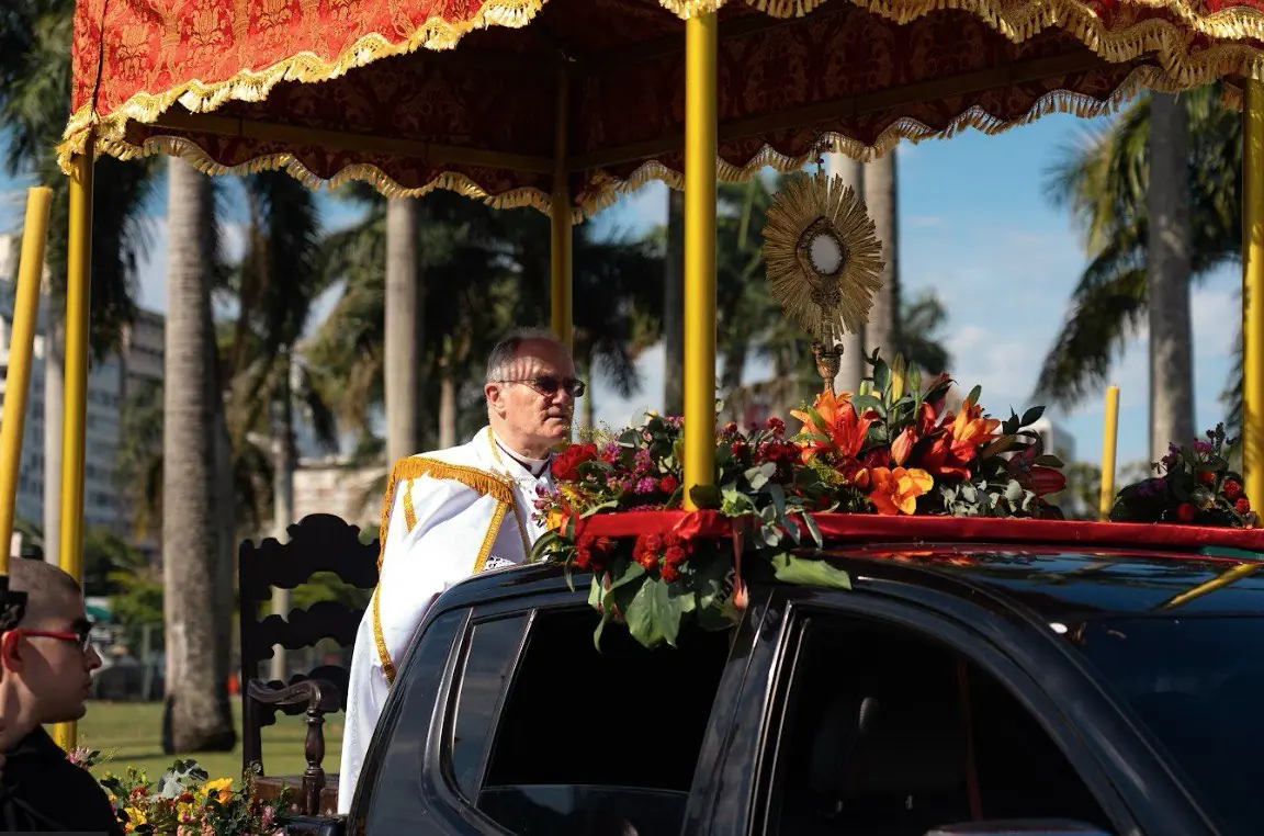 Dom Bernard Fellay na procissão solene de Cristo Rei, marcando o centenário da encíclica Quas Primas. Bispo Dom Bernard Fellay, em paramentos pontificais brancos e dourados, em uma procissão sob um dossel (baldaquino) vermelho. O ostensório do Santíssimo Sacramento está exposto sobre um carro ricamente florido, celebrando o centenário da festa de Cristo Rei instituída por Pio XI.