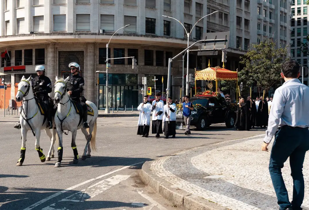 Cavalaria e procissão de Cristo Rei em frente ao Monumento do Santíssimo Sacramento, Centro do RJ. Vista de uma procissão religiosa com dois policiais montados a cavalo na vanguarda, seguido por clérigos e fiéis. Ao fundo, o carro com o Santíssimo Sacramento sob um dossel, próximo ao Monumento do Santíssimo Sacramento no centro do Rio de Janeiro. Prédios urbanos e palmeiras são visíveis.