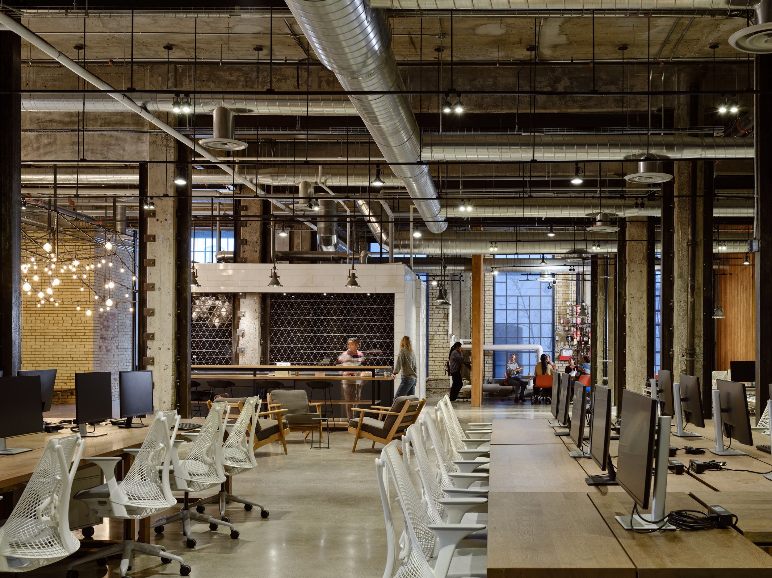 Modern industrial-style coworking space featuring exposed ductwork, communal workstations, and a collaborative lounge area with mixed seating arrangements.