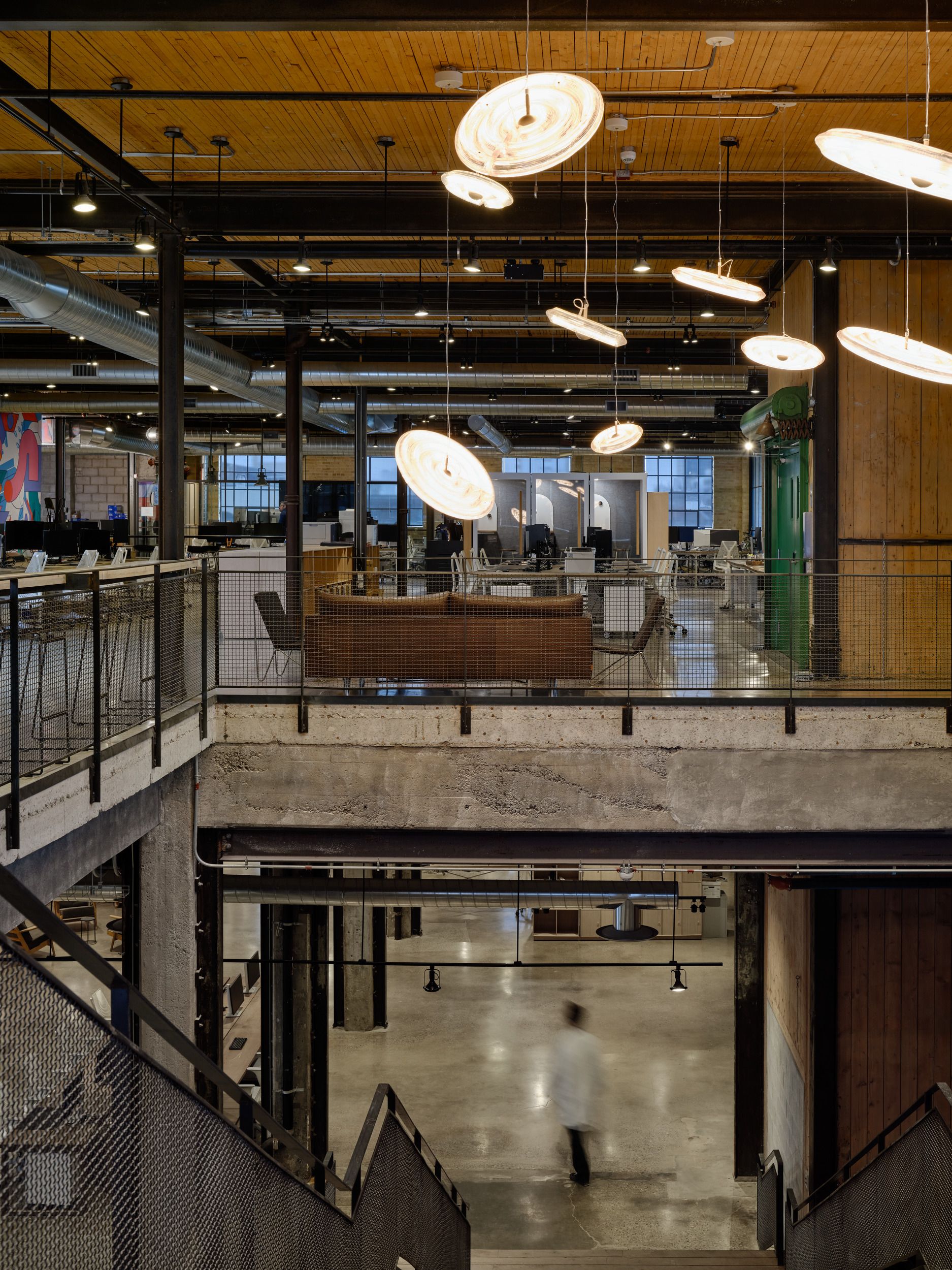 Modern industrial-style office space featuring an open two-level layout with exposed ceiling, distinctive circular pendant lighting, and concrete-wood architectural elements.