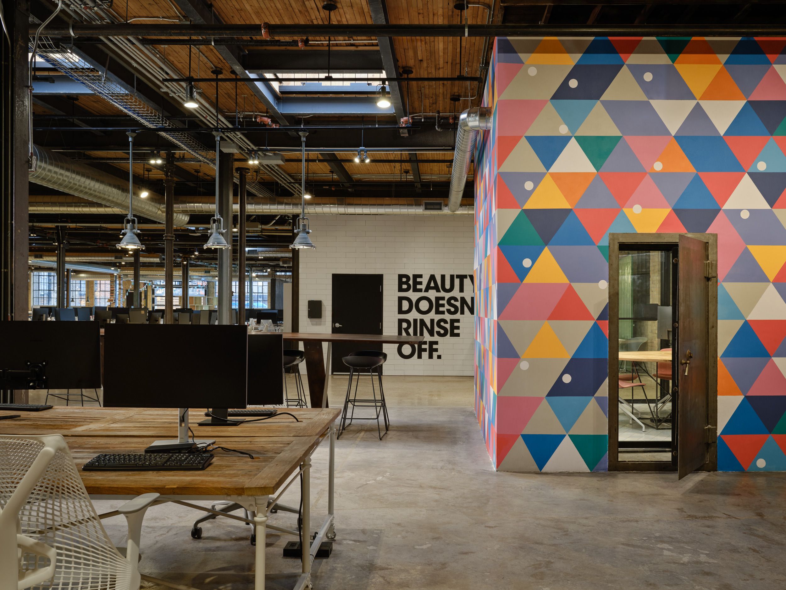 Modern office space featuring an industrial-style workspace with colorful geometric wall art and motivational text reading "Beauty Doesn't Rinse Off."