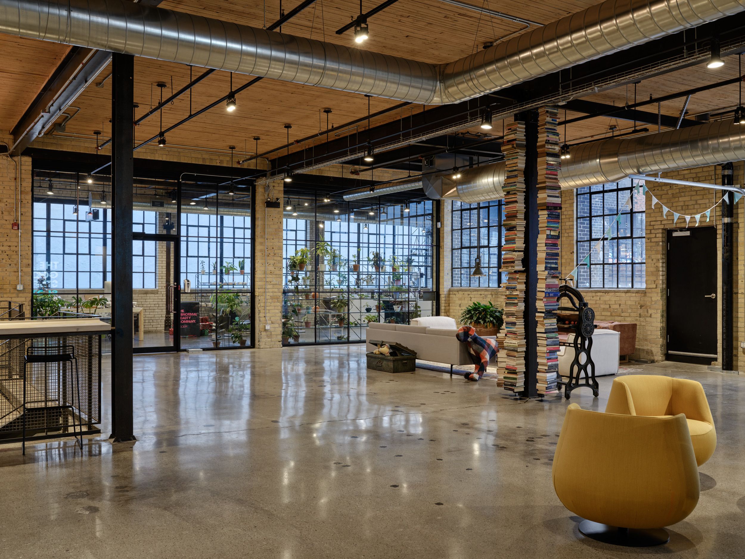 Modern industrial office space featuring exposed brick, large windows, concrete floors, and casual seating areas with plants and contemporary furnishings.