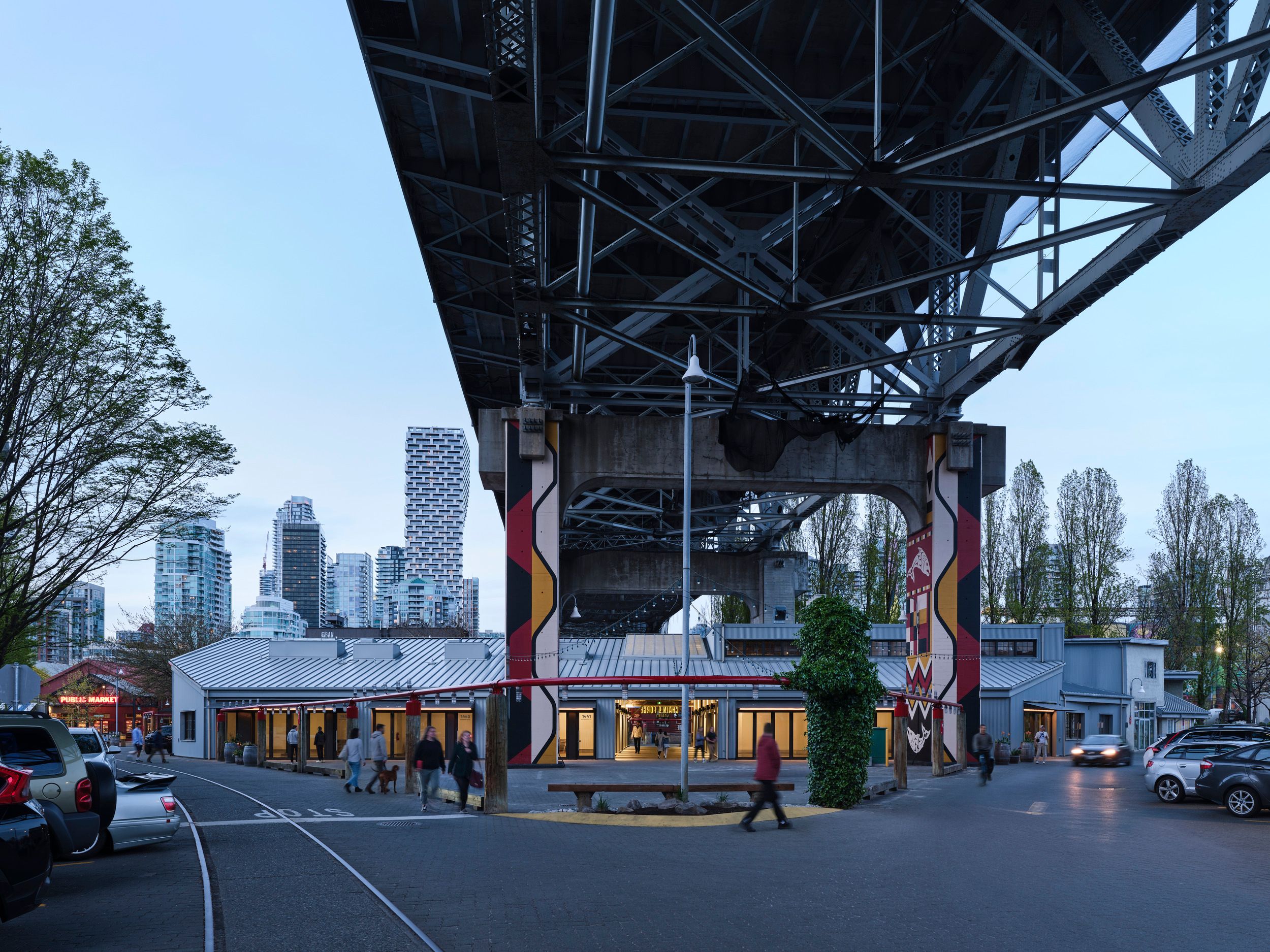View of Granville Island marketplace buildings beneath the Granville Bridge, with Vancouver's downtown skyline visible in the background.