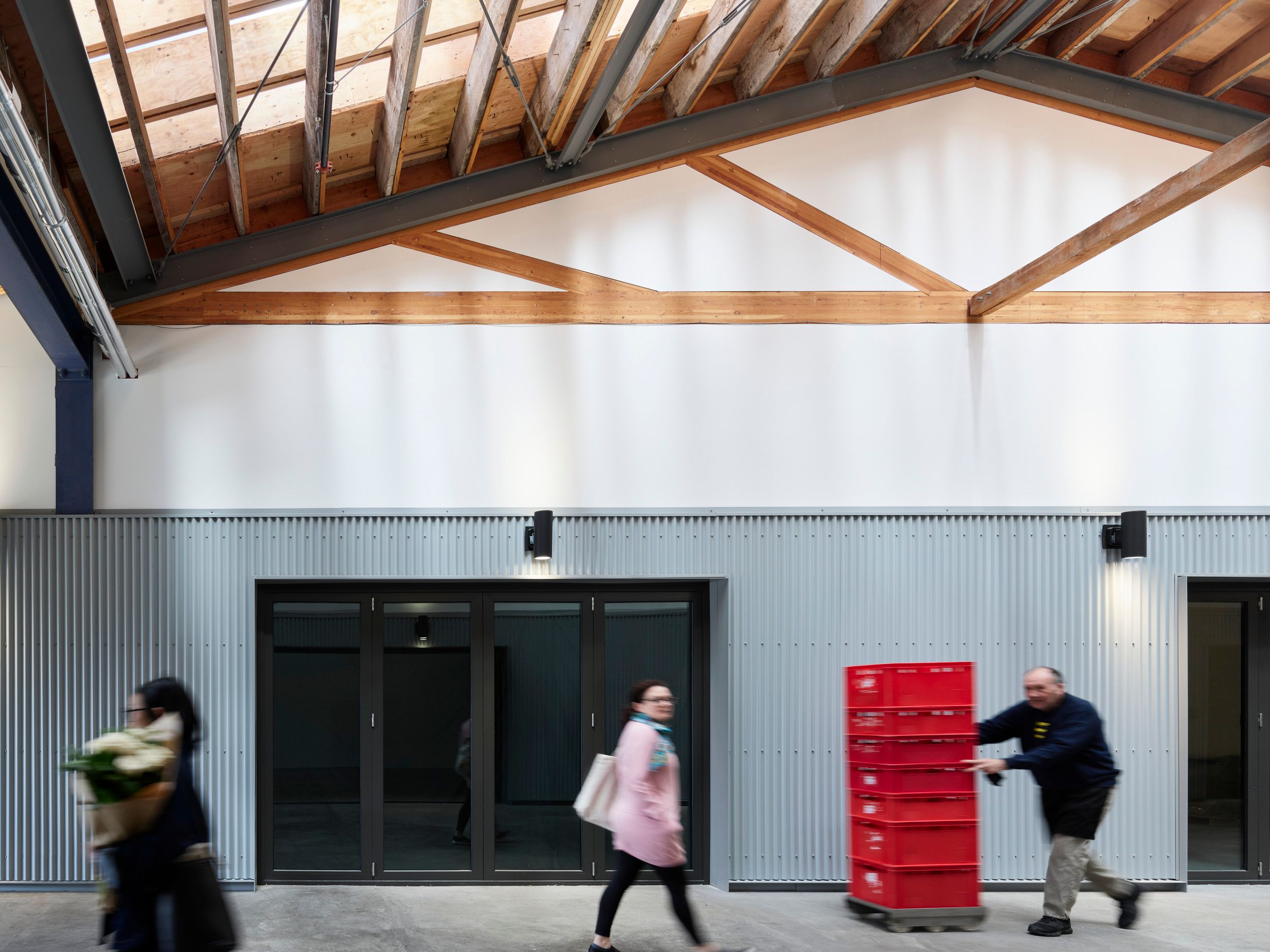 Interior warehouse corridor with exposed wooden trusses, corrugated metal walls, and glass doors showing daily workplace activity.