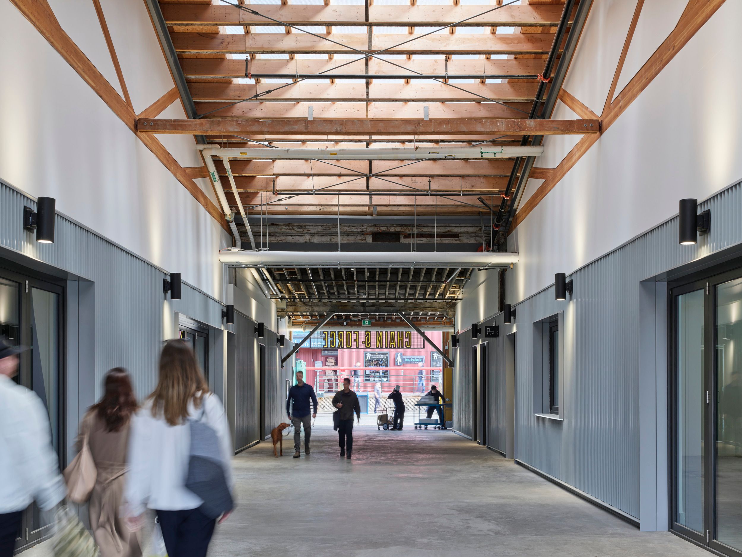 Interior walkway of a modern commercial building featuring exposed wooden ceiling beams, grey walls, and a covered passageway leading to an outdoor area.