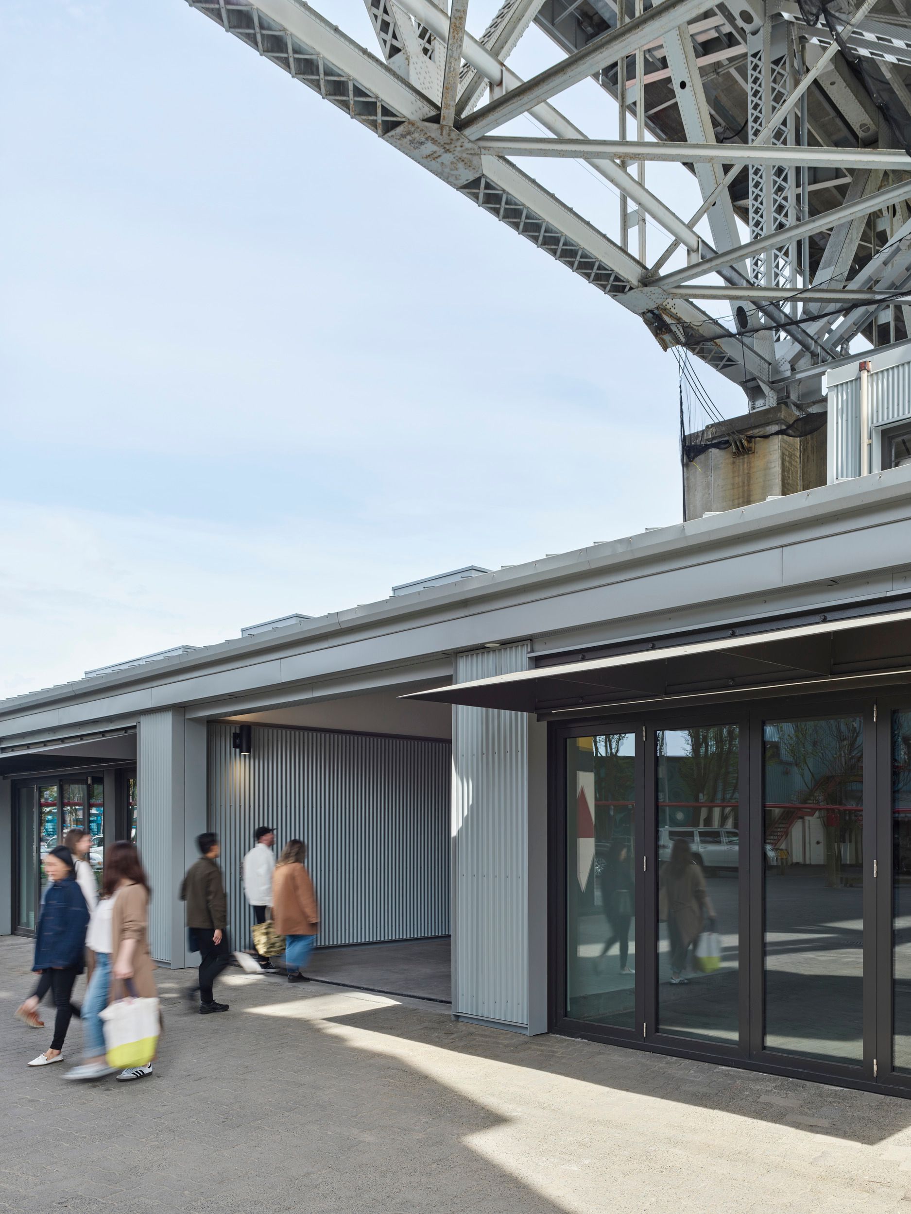 Modern space underneath a bridge or elevated structure, featuring glass storefronts and corrugated metal finishes.