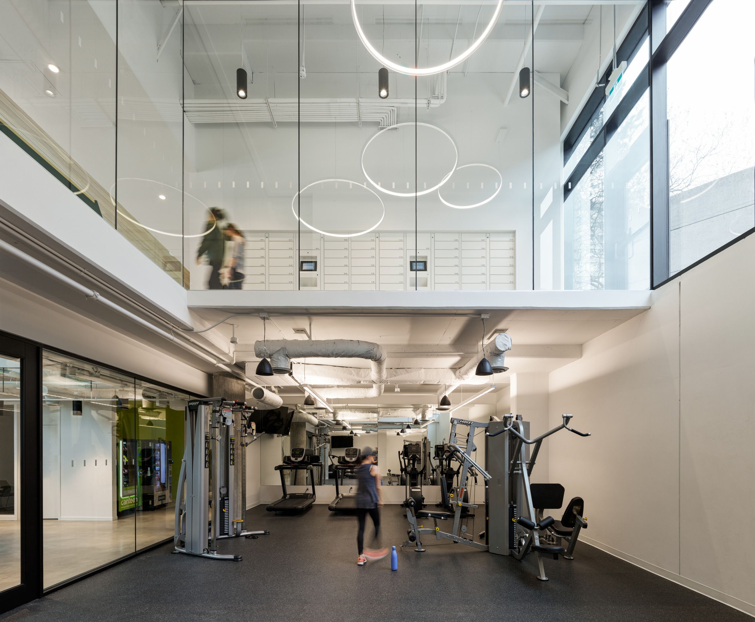 Modern two-level fitness facility featuring exercise equipment on the ground floor and mail storage lockers visible through glass railings on the mezzanine level, with contemporary circular lighting fixtures and floor-to-ceiling windows.