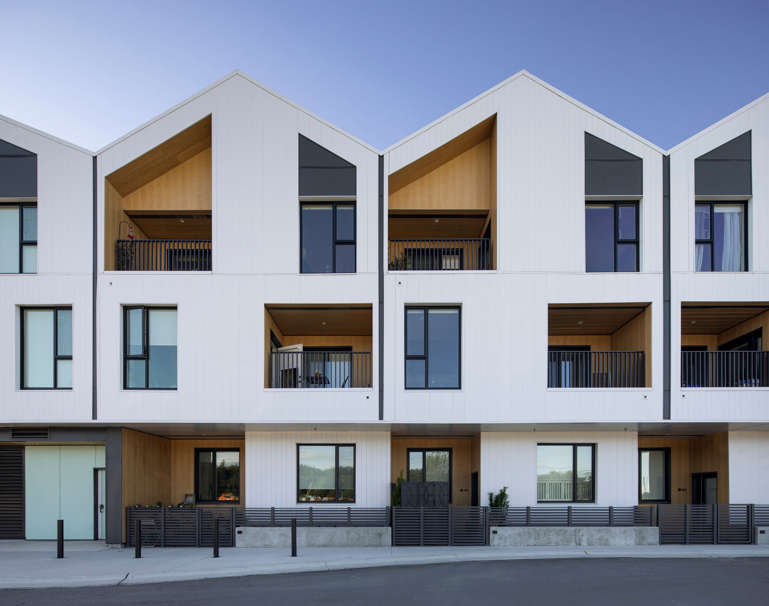 Modern townhouse development featuring white gabled facades with three levels, recessed balconies, and contemporary black metal railings.