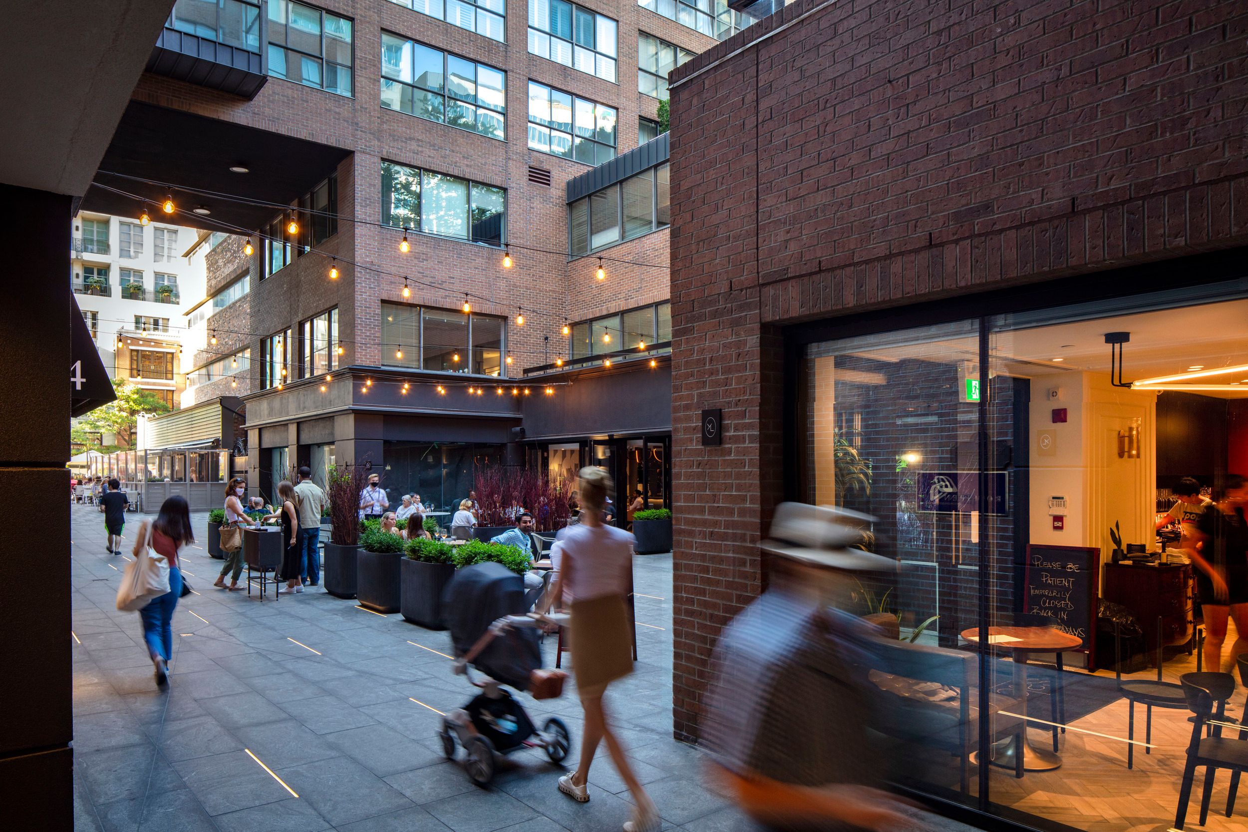 Modern urban courtyard with outdoor café seating, string lighting, and retail spaces along a pedestrian walkway between brick buildings.