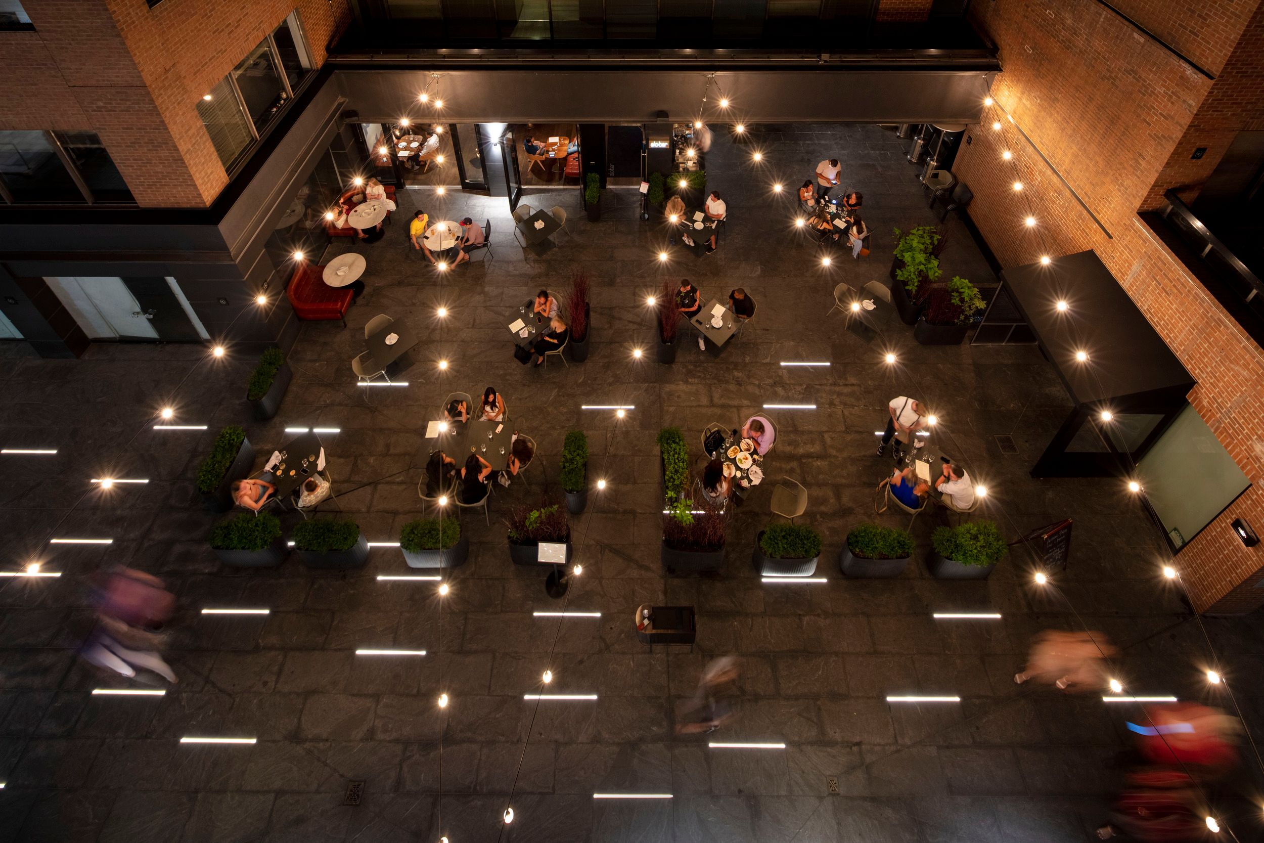 Aerial view of an outdoor dining courtyard at night with string lights, spaced tables, and illuminated floor patterns.