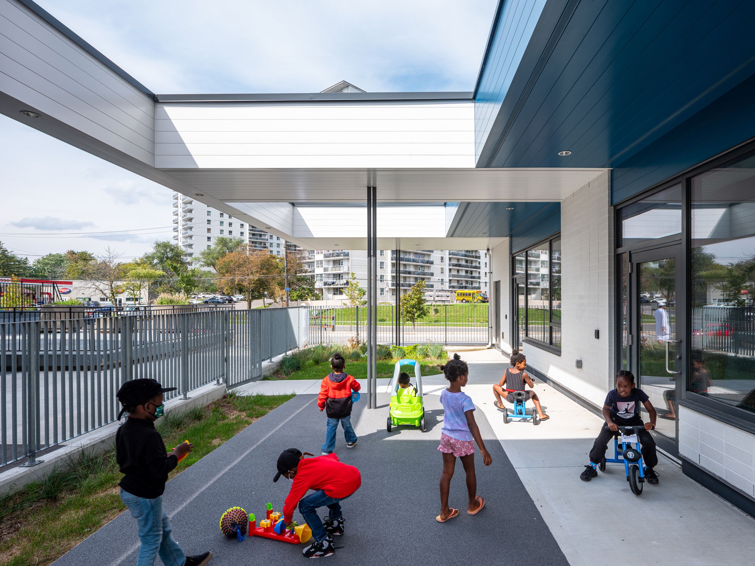 Covered outdoor play area at a childcare facility with riding toys and play equipment, featuring modern architecture and residential buildings visible in the background.