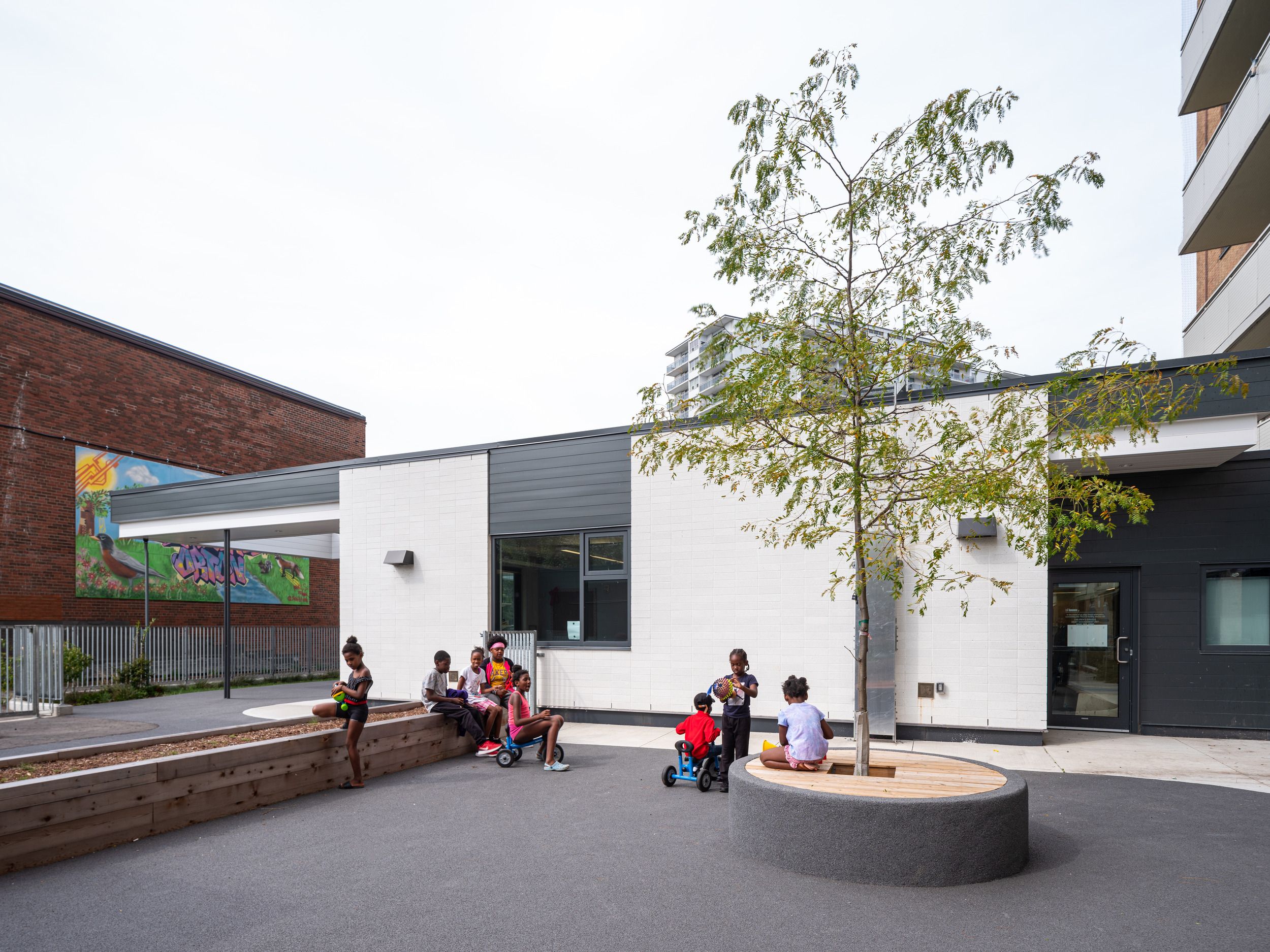 Modern childcare facility courtyard featuring a wooden bench seating area, planted tree, and colorful mural on the exterior wall.