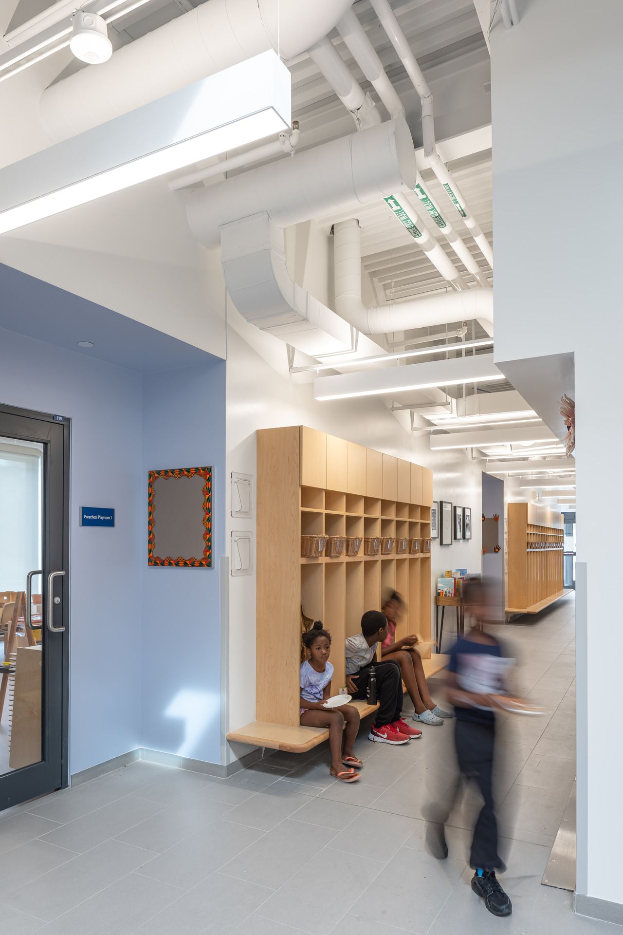 School hallway with built-in wooden cubbies and benches showing exposed utility systems on white ceiling.