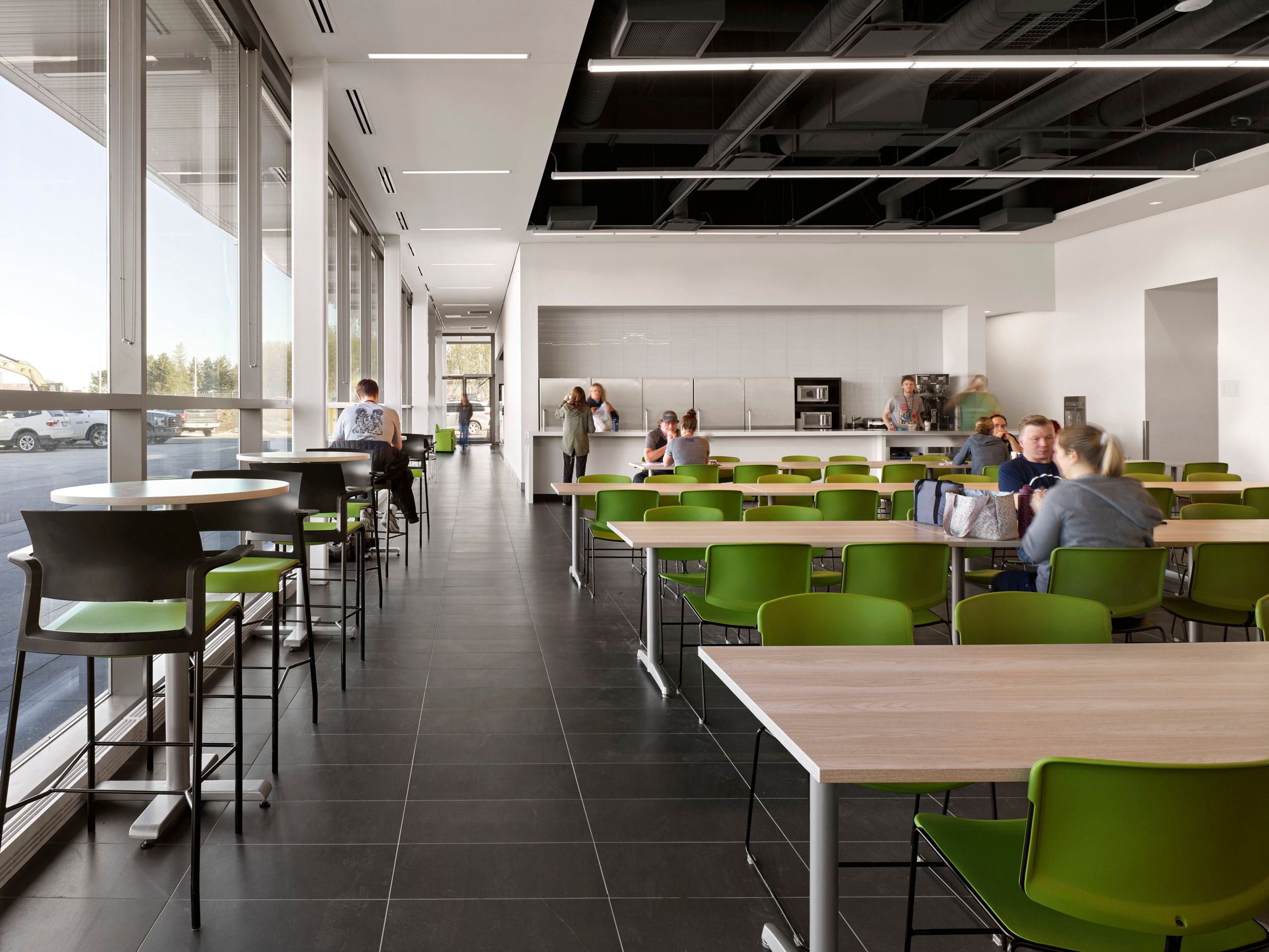 Modern cafeteria space featuring bright green seating, floor-to-ceiling windows, and a serving counter with kitchen area in the background.
