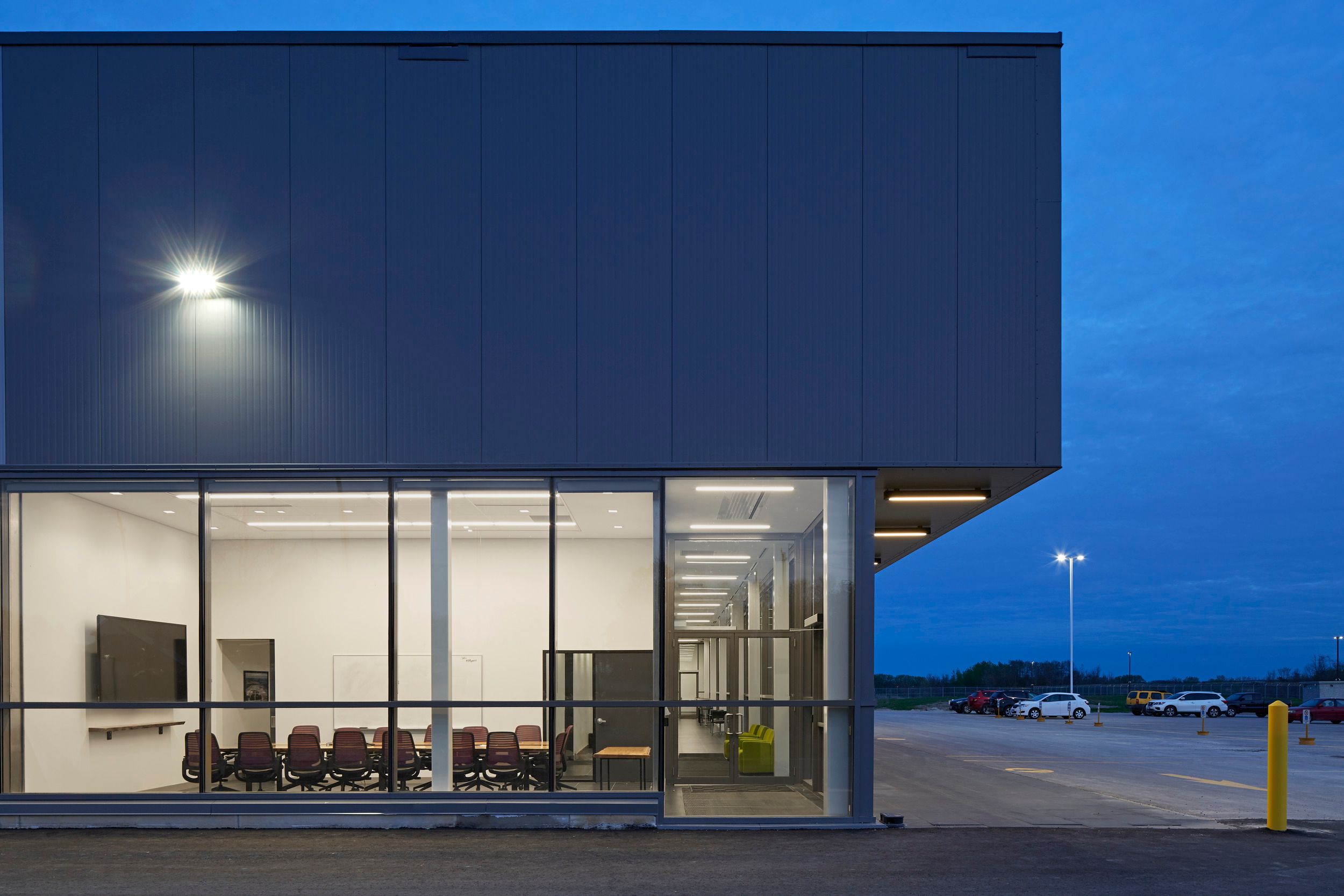 Modern commercial building exterior with illuminated conference room visible through floor-to-ceiling windows at dusk, adjacent to parking lot.