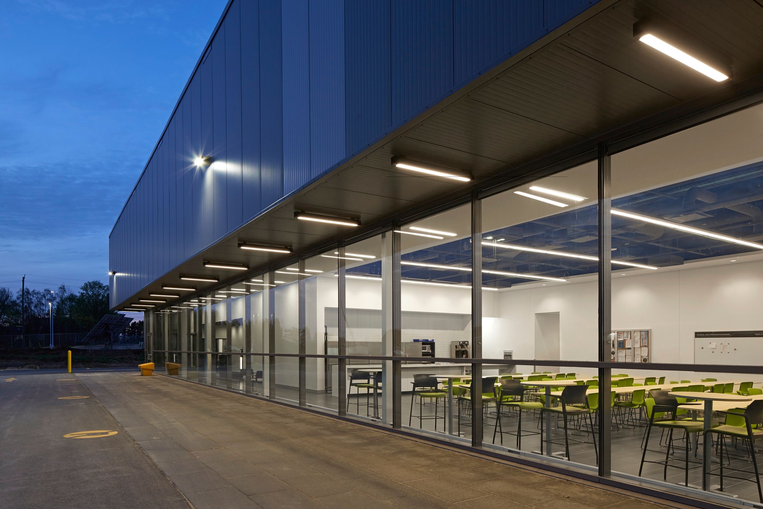 Modern industrial building exterior with floor-to-ceiling windows showing a brightly lit cafeteria or break room with lime green seating at dusk.