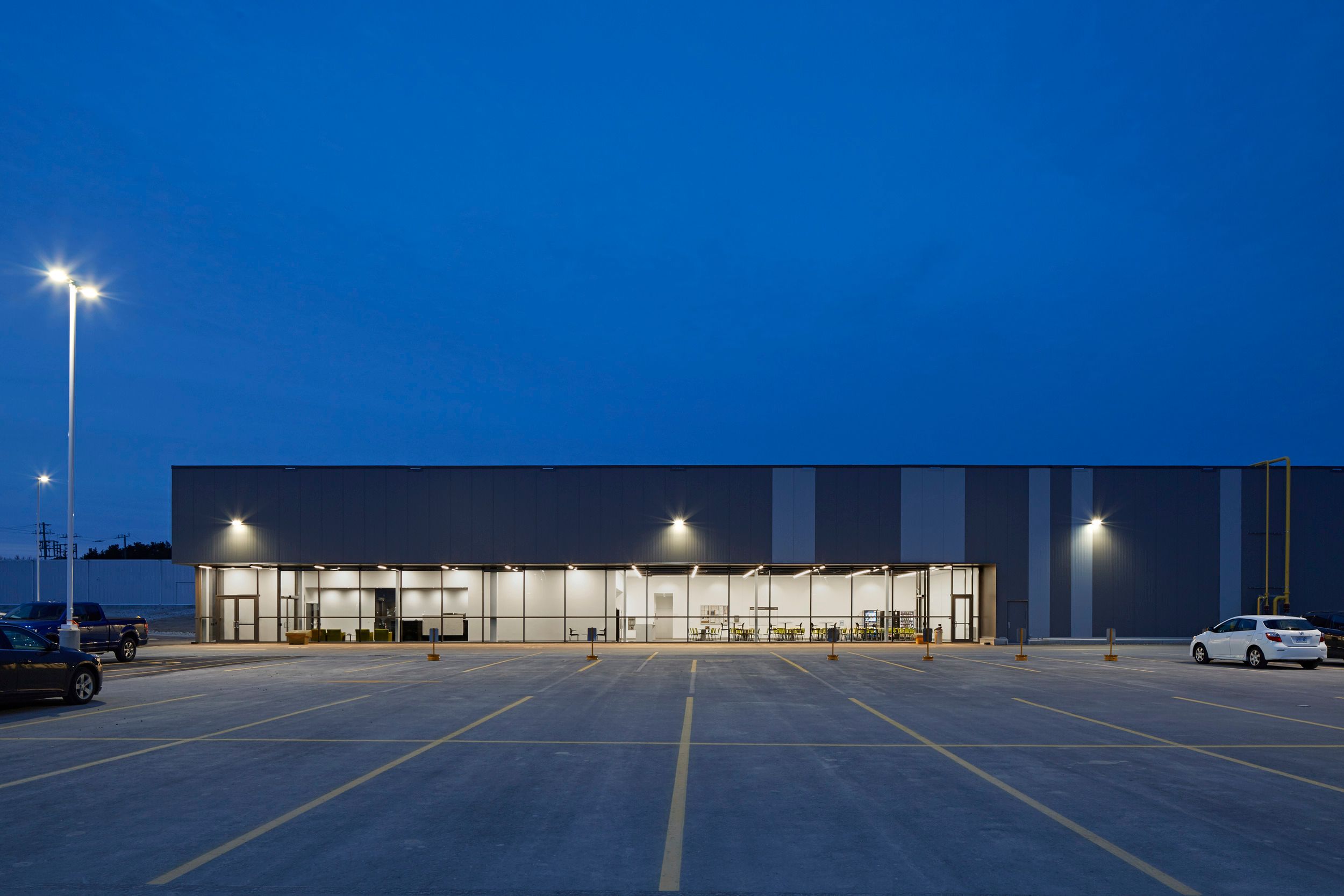 Exterior view of a modern commercial building with illuminated storefronts and a spacious parking lot at dusk.