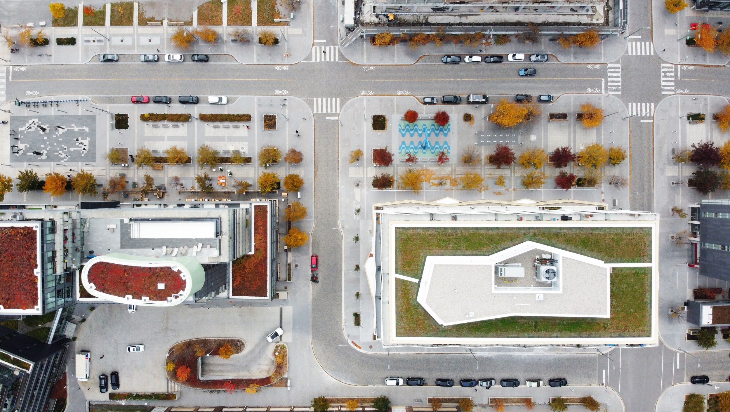 Aerial view of an urban development featuring modern buildings with distinctive rooftops, parking areas, and autumn-colored street trees.