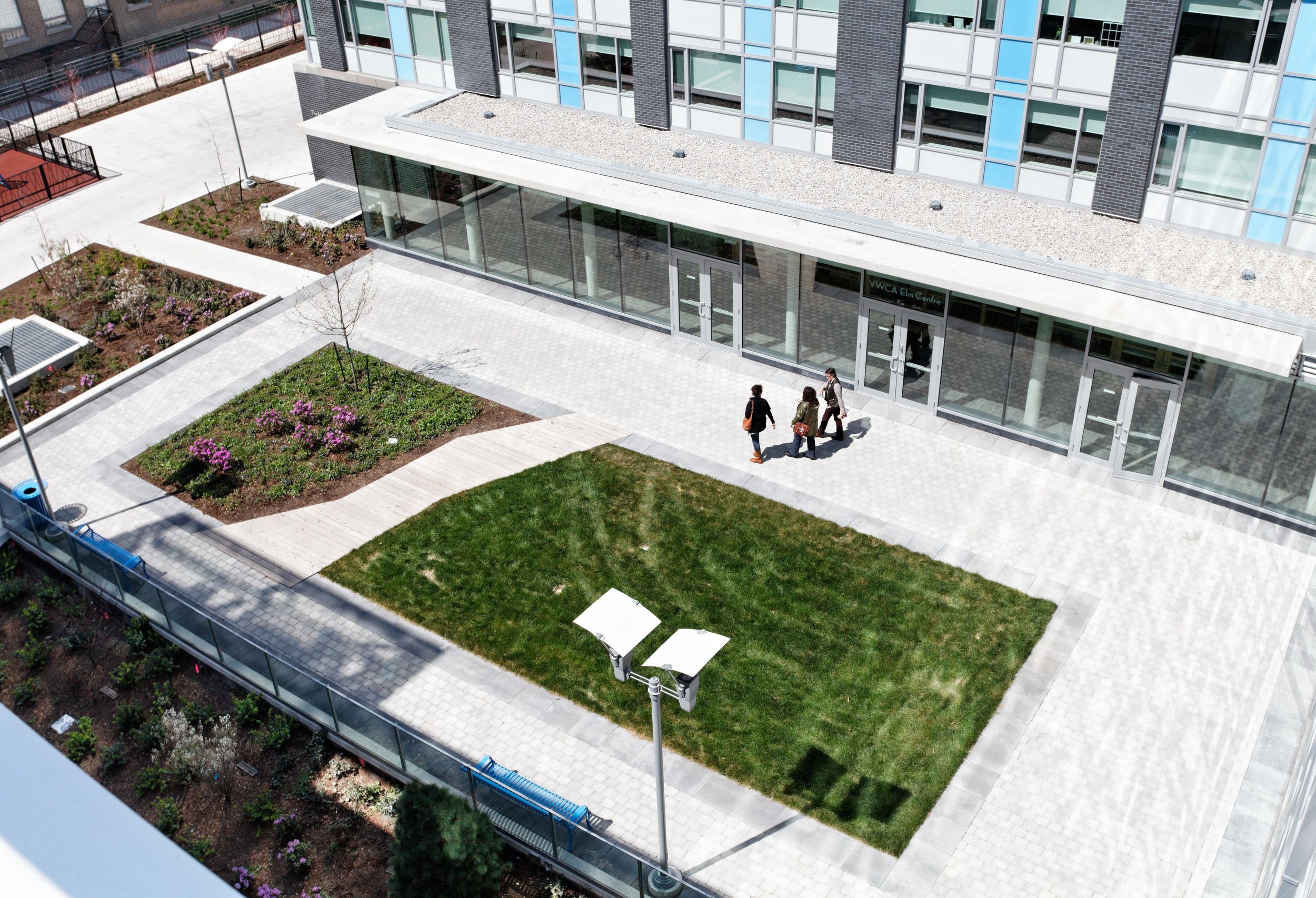 Modern building entrance with landscaped courtyard featuring grass areas, planted beds, and concrete walkways viewed from above.