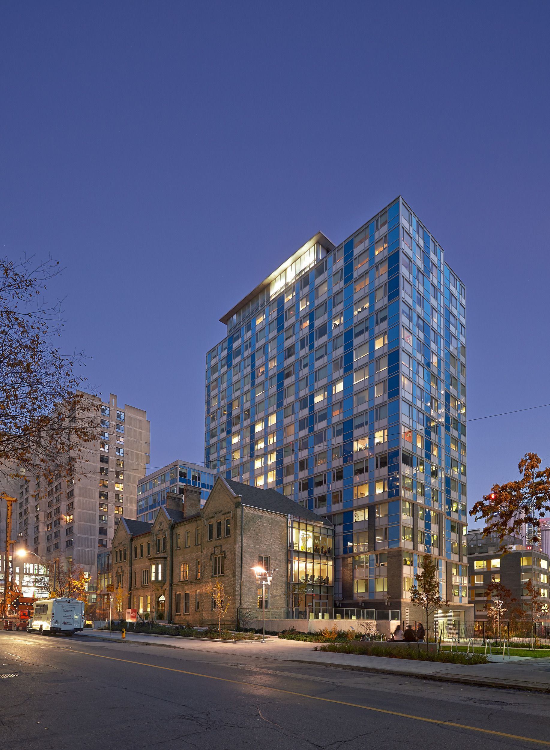 Modern glass high-rise residential tower integrated with preserved historic stone buildings at street level in an urban setting at dusk.